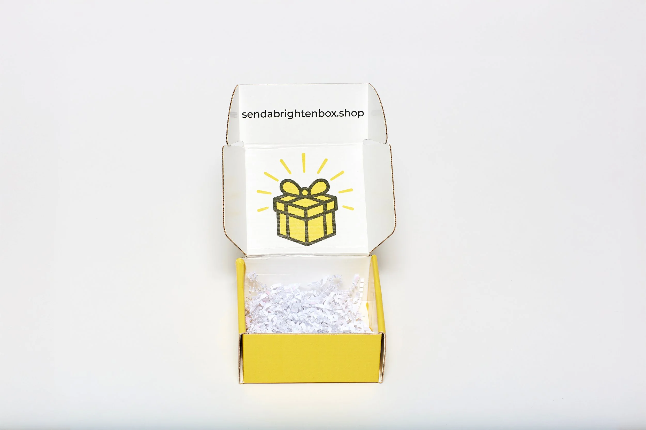 Open yellow gift box with white shredded paper inside, illustration of a yellow gift box with a bow on the inside of the lid, and the URL 'sendabrightenbox.shop' on the interior flap.