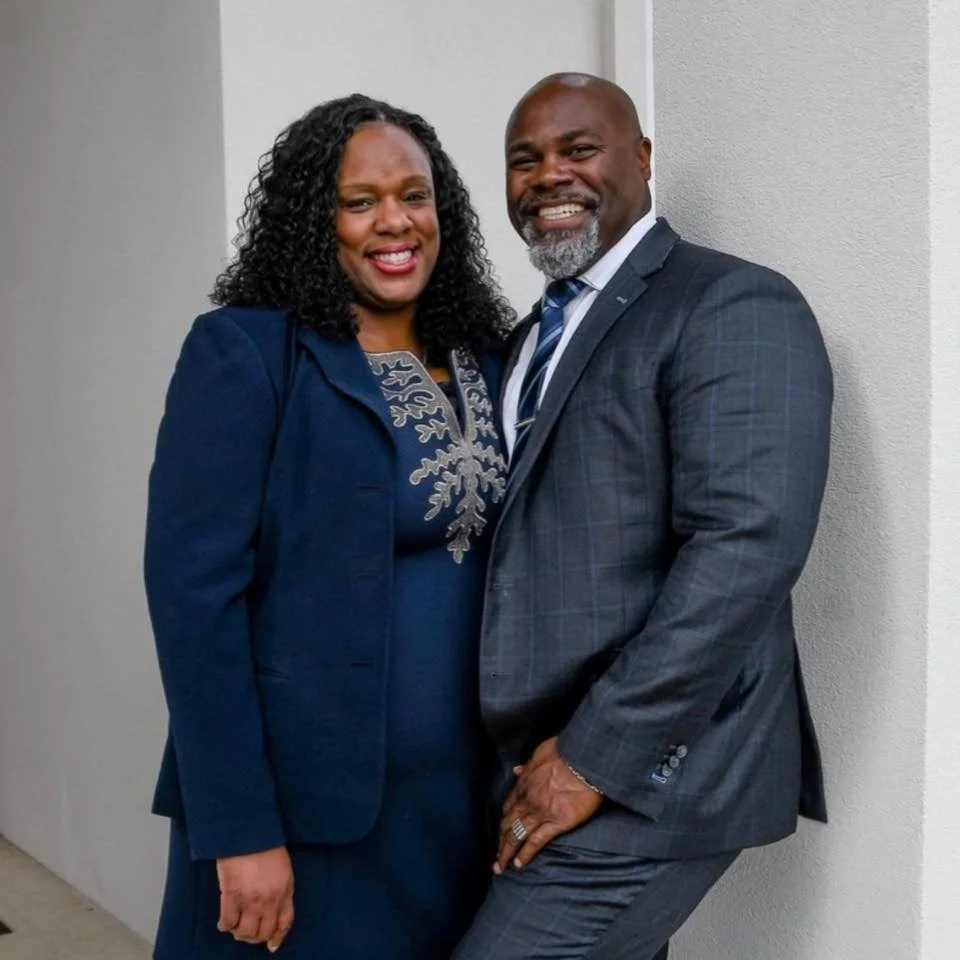 A smiling African American couple standing close together, dressed in formal business attire, against a plain light-colored wall.