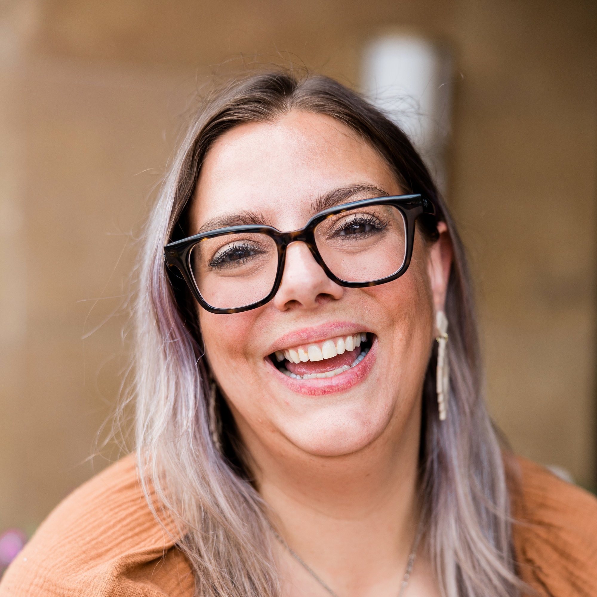 A woman smiling and wearing glasses with a blurred outdoor background.