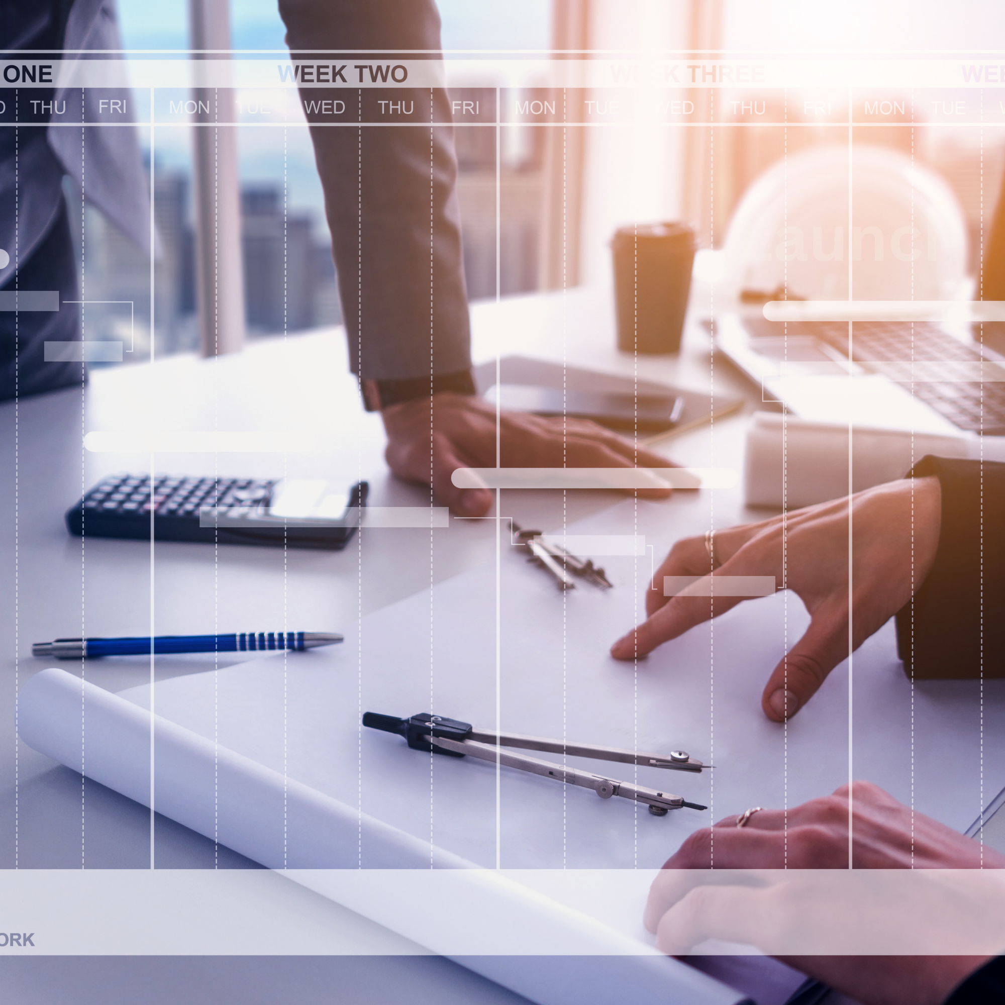 People working on a business project with a blueprint, calculator, pen, compass, and coffee on a desk in a modern office.