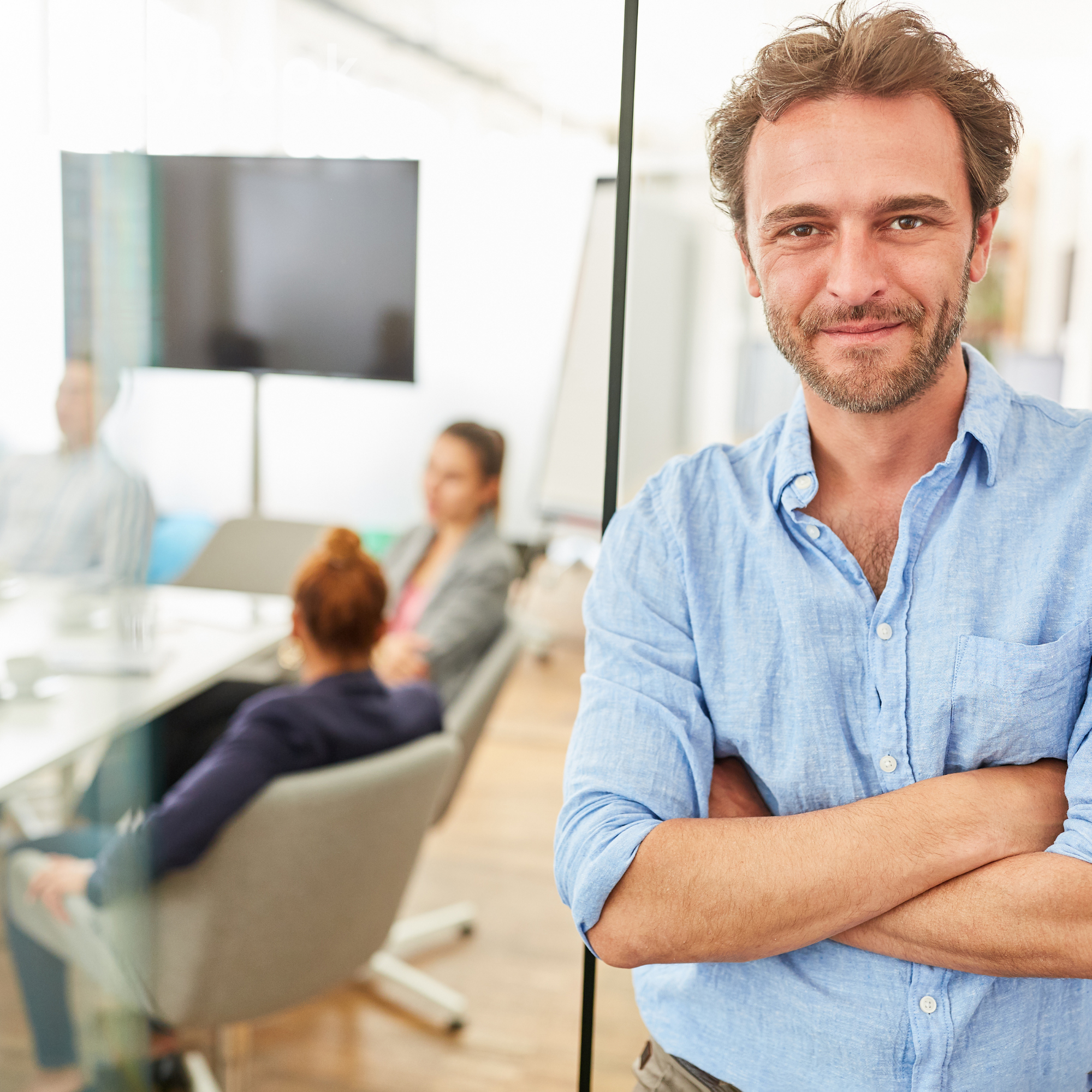 A man with curly hair and a beard smiling with crossed arms in a bright, modern office conference room, with blurred colleagues sitting at a table in the background.