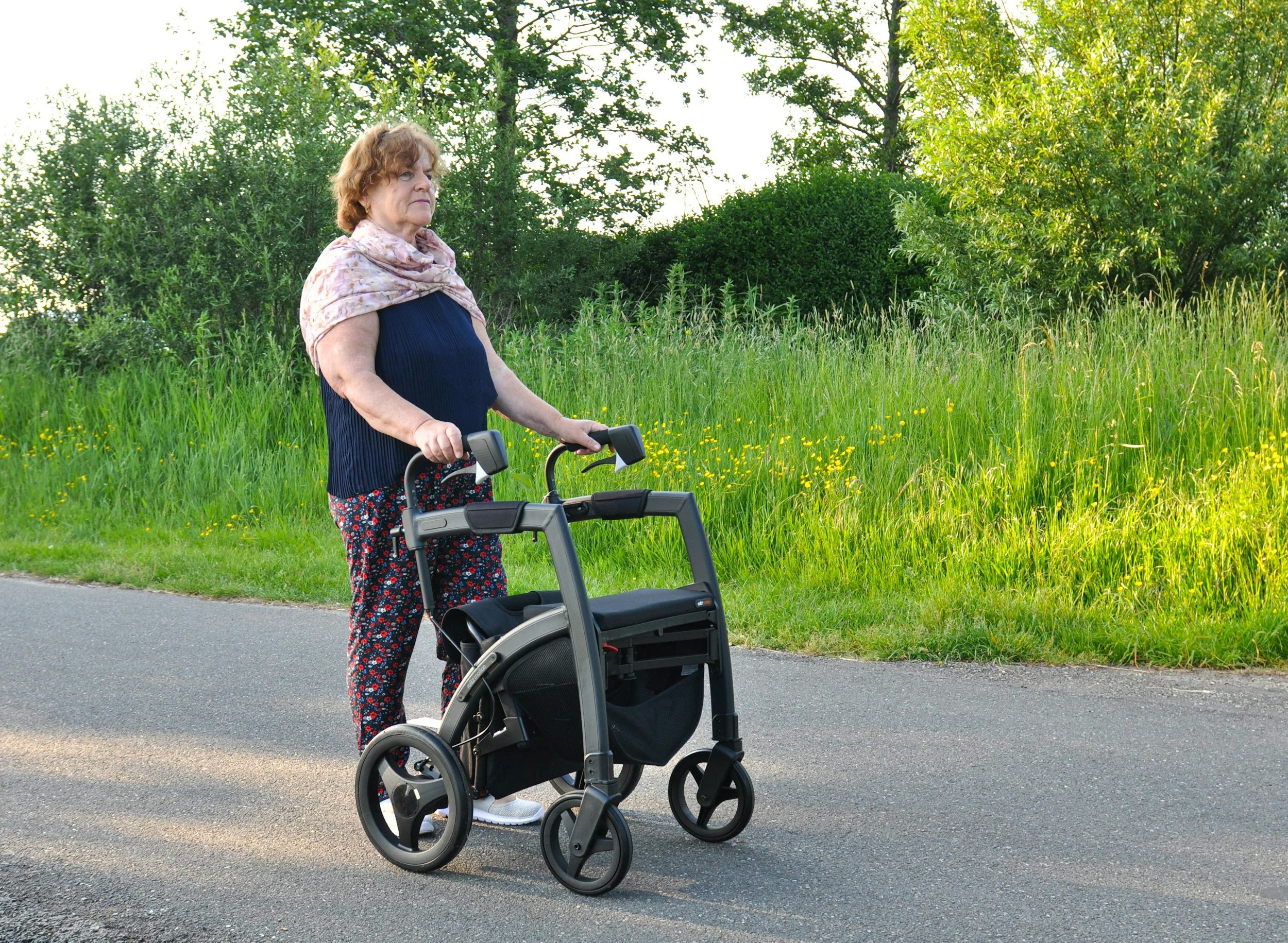An elderly woman walking with a walker along a paved path in a park during daytime, with green grass, yellow flowers, trees, and bushes in the background.