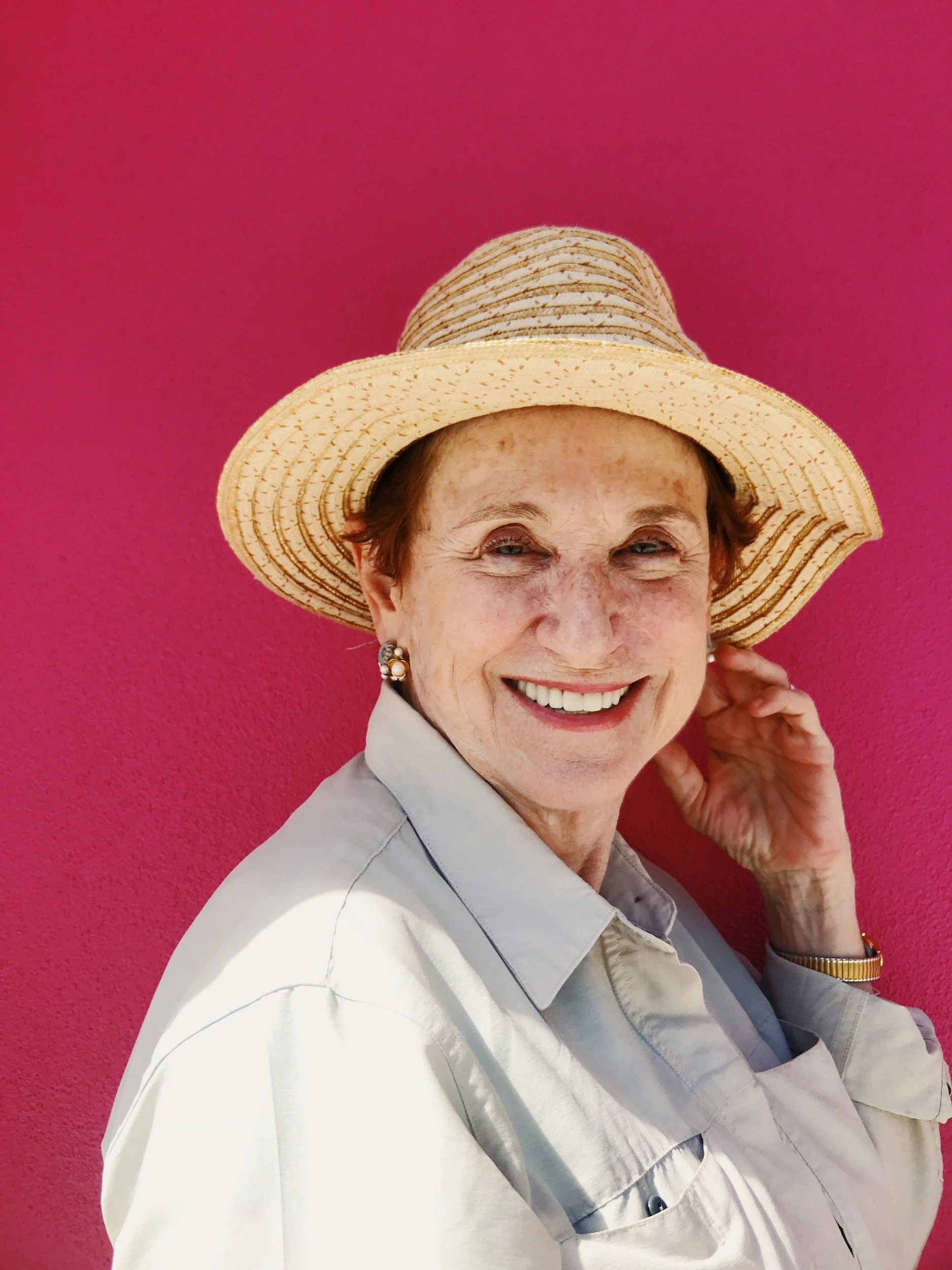 Smiling elderly woman wearing a wide-brimmed straw hat, earrings, and a light-colored shirt, standing against a bright pink wall.