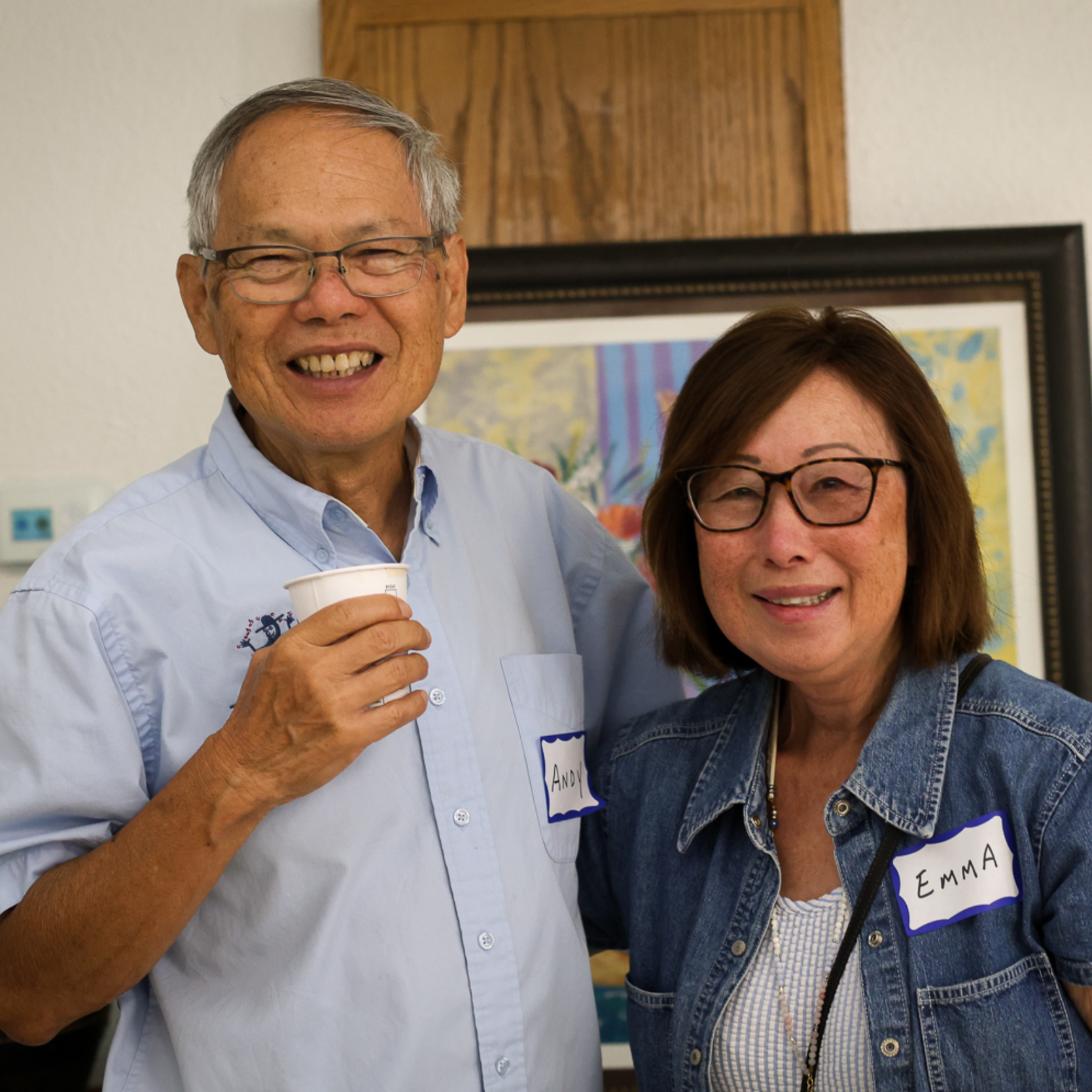 A man and woman smiling, standing close together at a social event, each wearing a name tag, with the man holding a small cup.