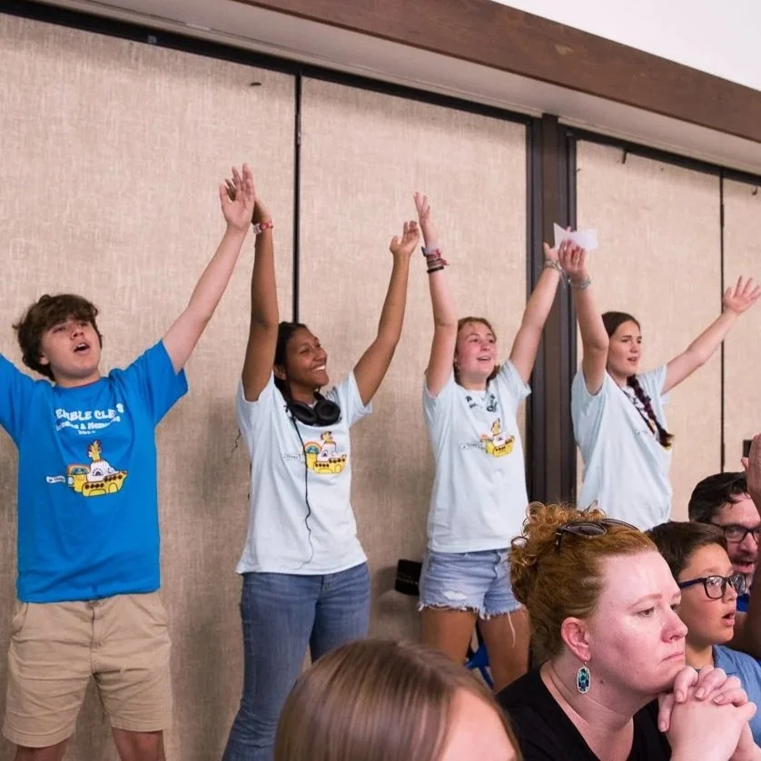 Four young people standing in a row with their arms raised, smiling and cheering. Two women and two men, all wearing white or blue T-shirts featuring a yellow vehicle and the word 'Goble'. They are inside a room with beige wall panels.