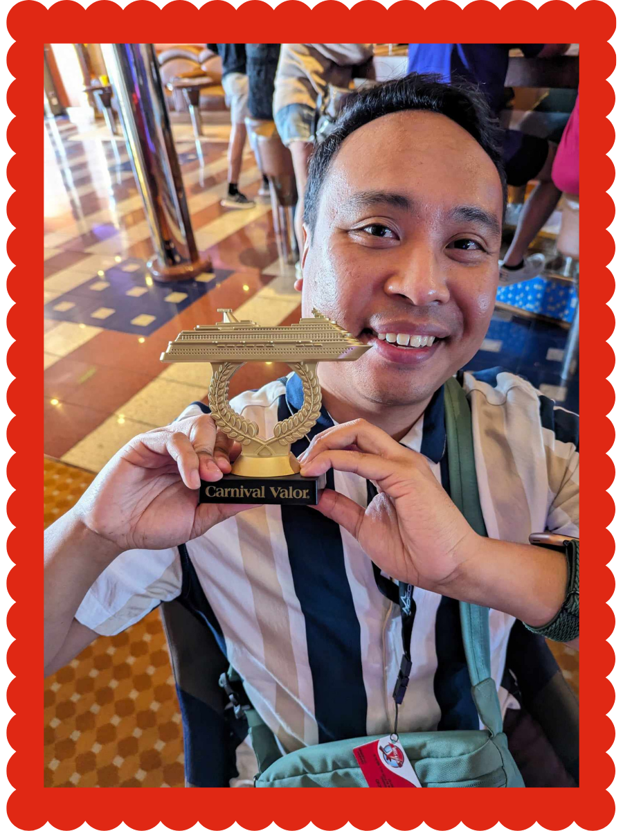 A man smiling and holding a trophy with a cruise ship on it and the words "Carnival Valor" at a cruise terminal or lobby.