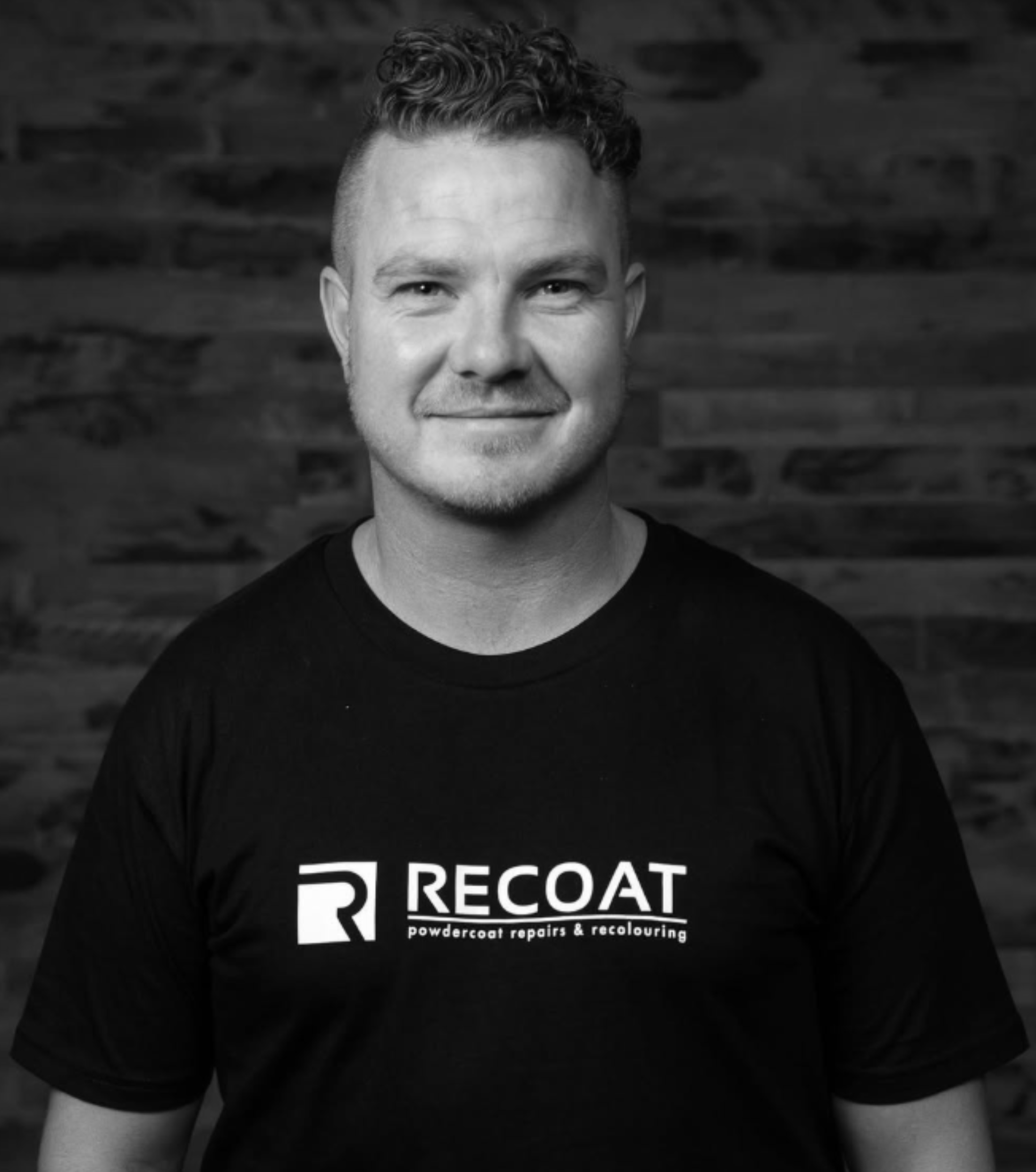 Close-up of man with curly hair wearing a black t-shirt with a logo for Recoat powdercoat repairs and recolouring, standing against a wooden background.