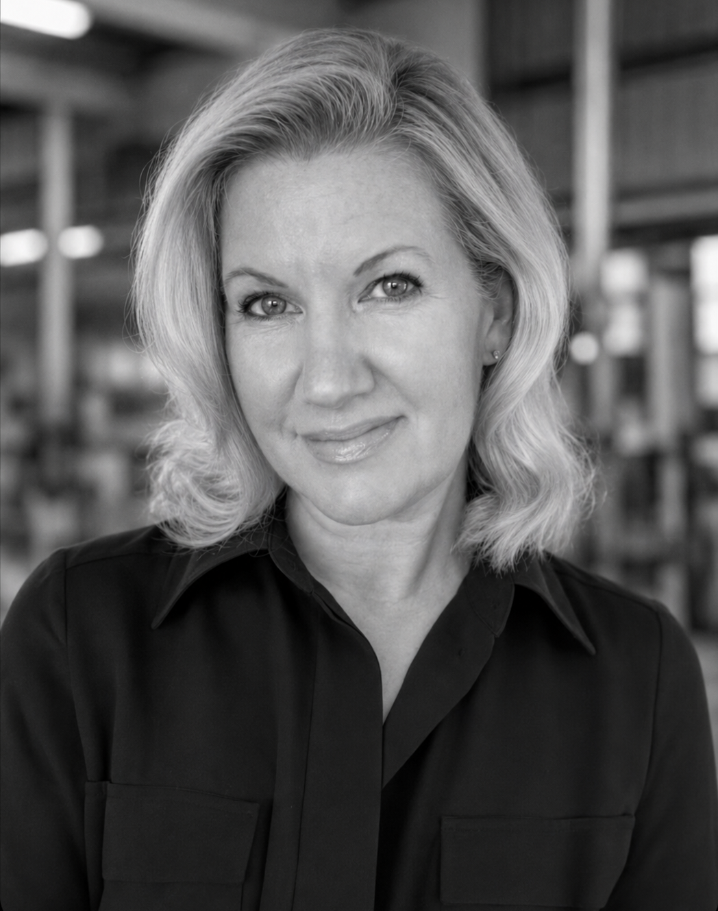 Black and white headshot of a woman with wavy blonde hair, wearing a dark collared shirt, indoors with blurred shelves in the background.