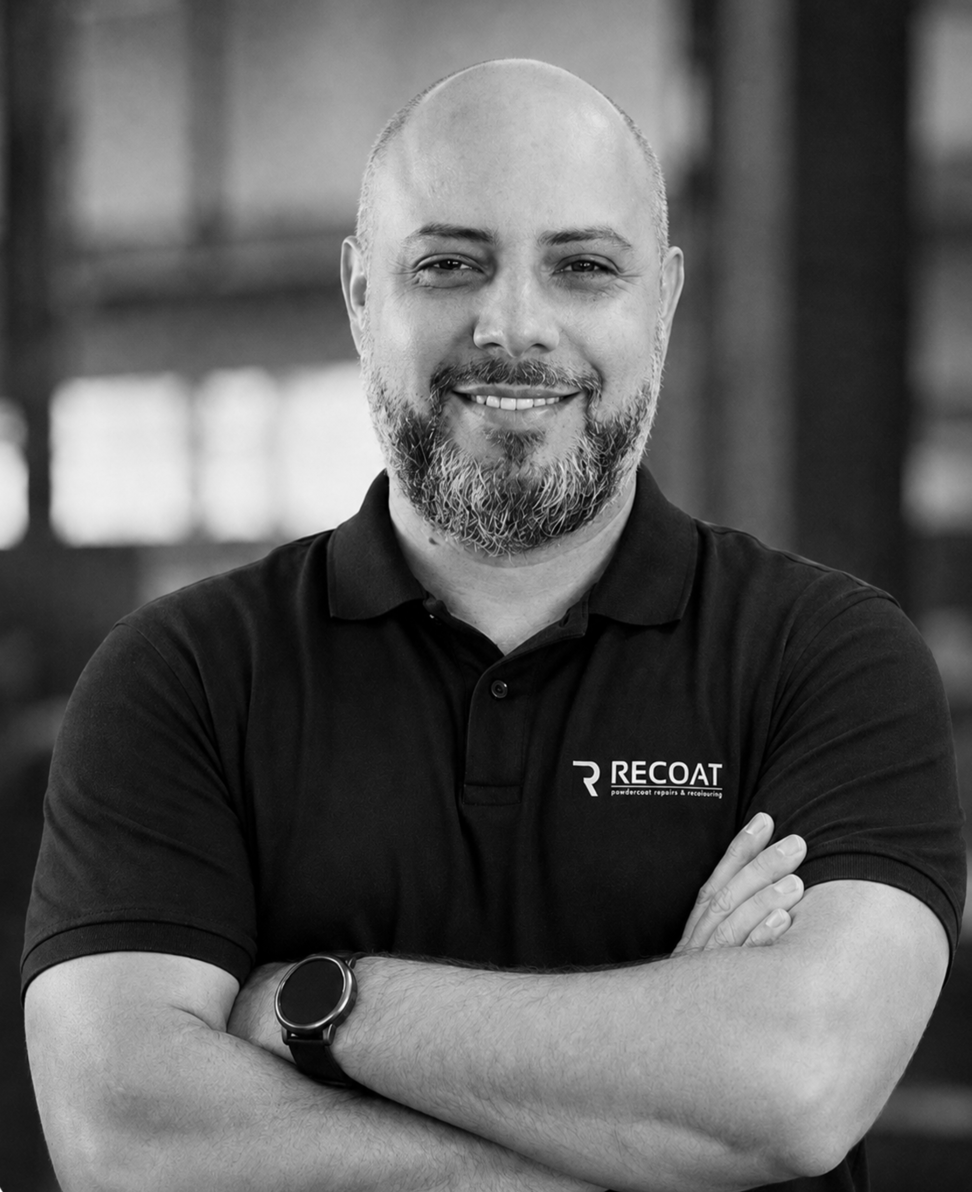Close-up of a smiling man with a beard, wearing a black polo shirt with a company logo, arms crossed, in an indoor setting.