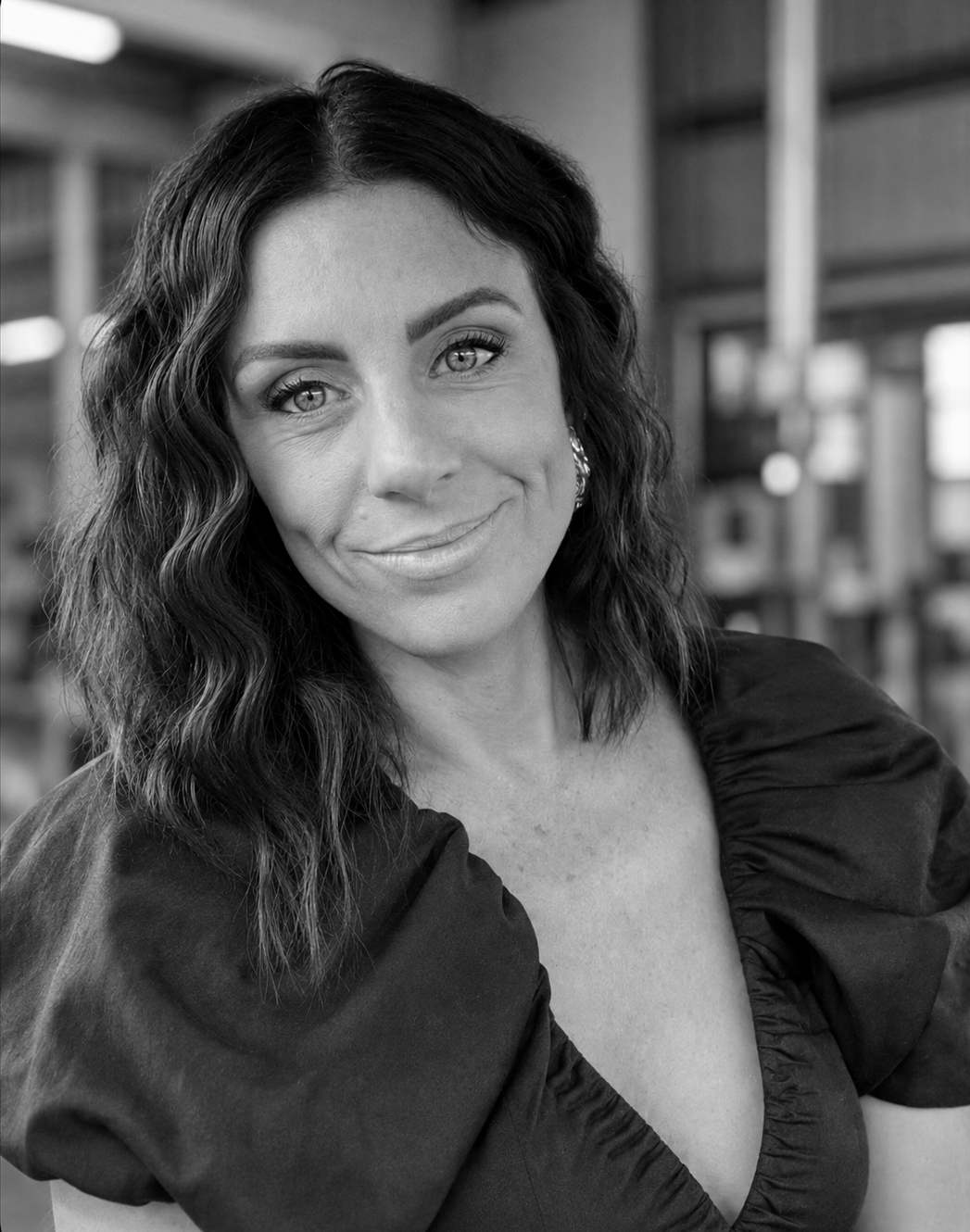 Black and white portrait of a woman with wavy hair, smiling, wearing earrings, in front of a blurred indoor background.