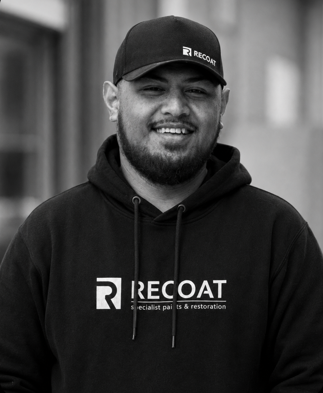 A smiling man with facial hair and a hat, wearing a hoodie with the logo and words "RECOAT specialist paints & restoration". Black and white photo outdoors.