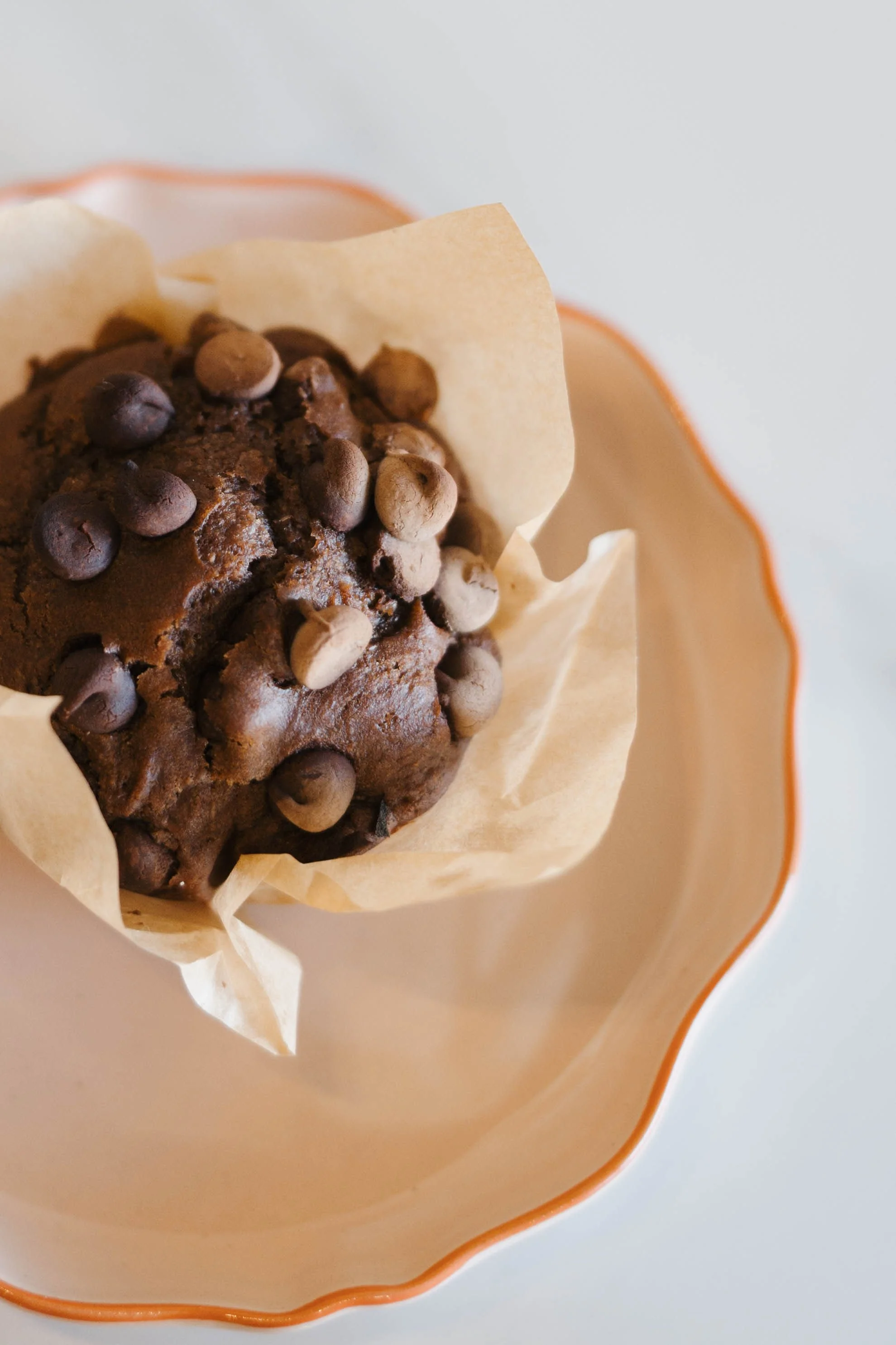 Chocolate muffin with chocolate chips in baking paper liner, inside a beige bowl with orange rim.