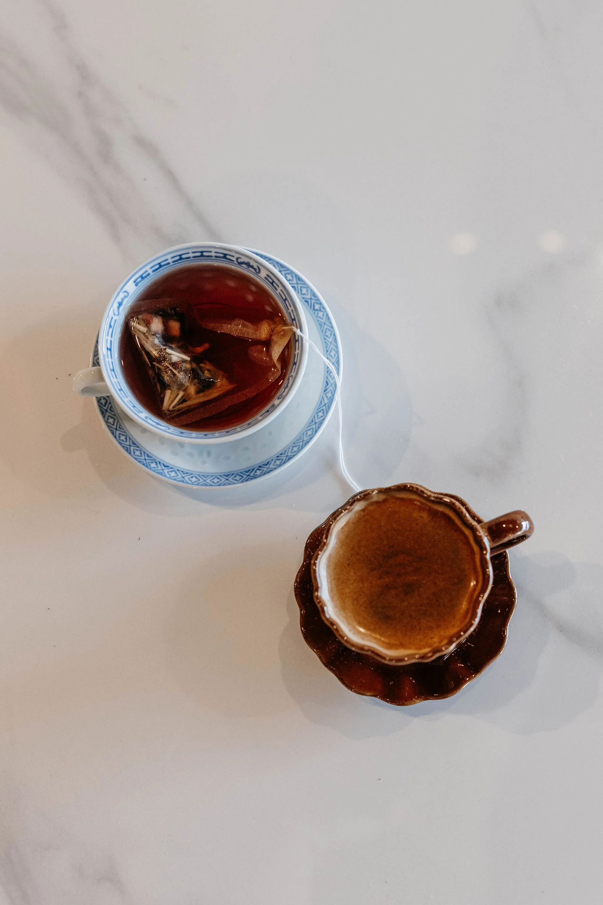A cup of tea with tea bags inside, placed on a small matching saucer, and a brown coffee cup with a beverage, on a white marble surface.