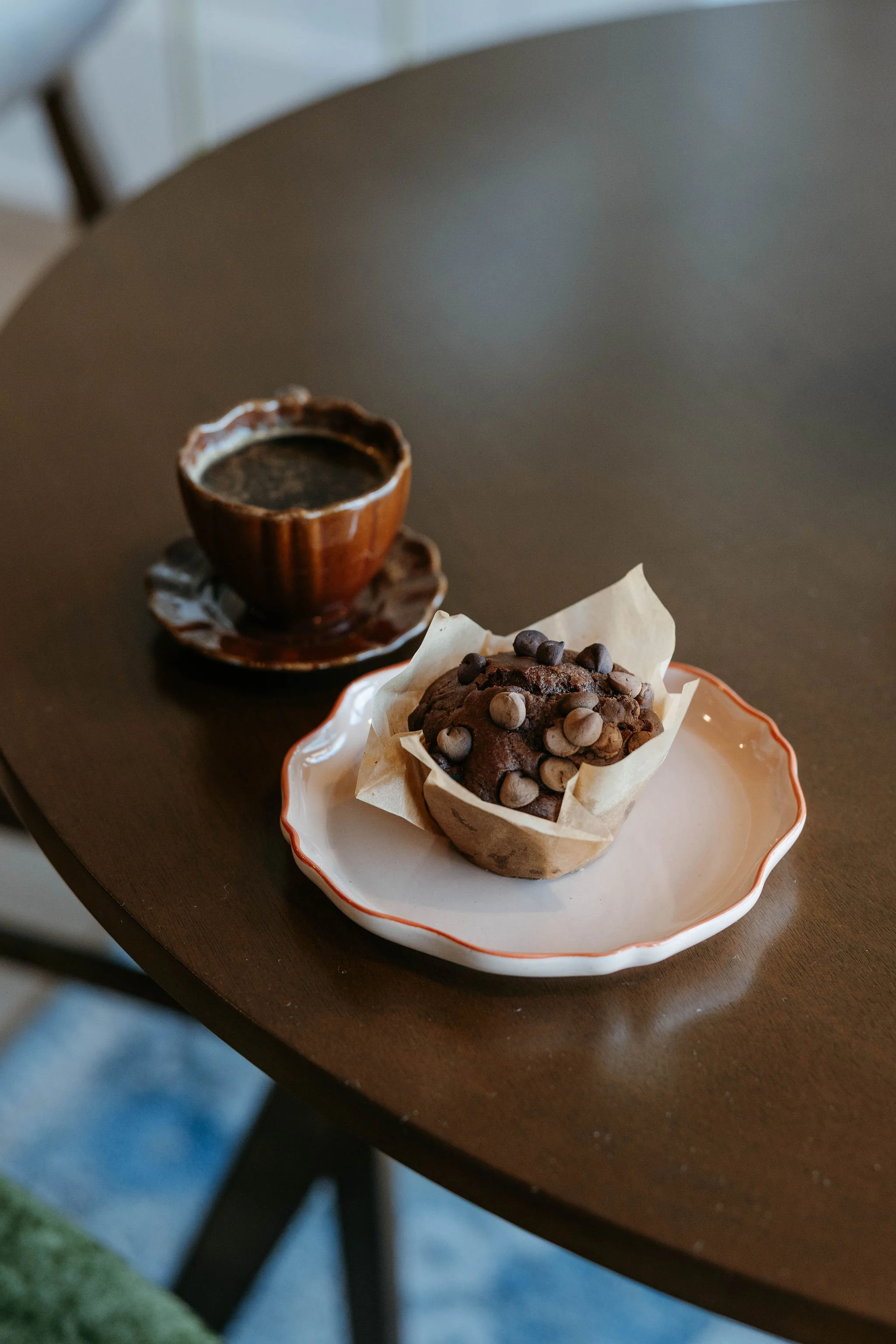 A chocolate muffin with chocolate chips on a white plate and a cup of coffee on a wooden table.