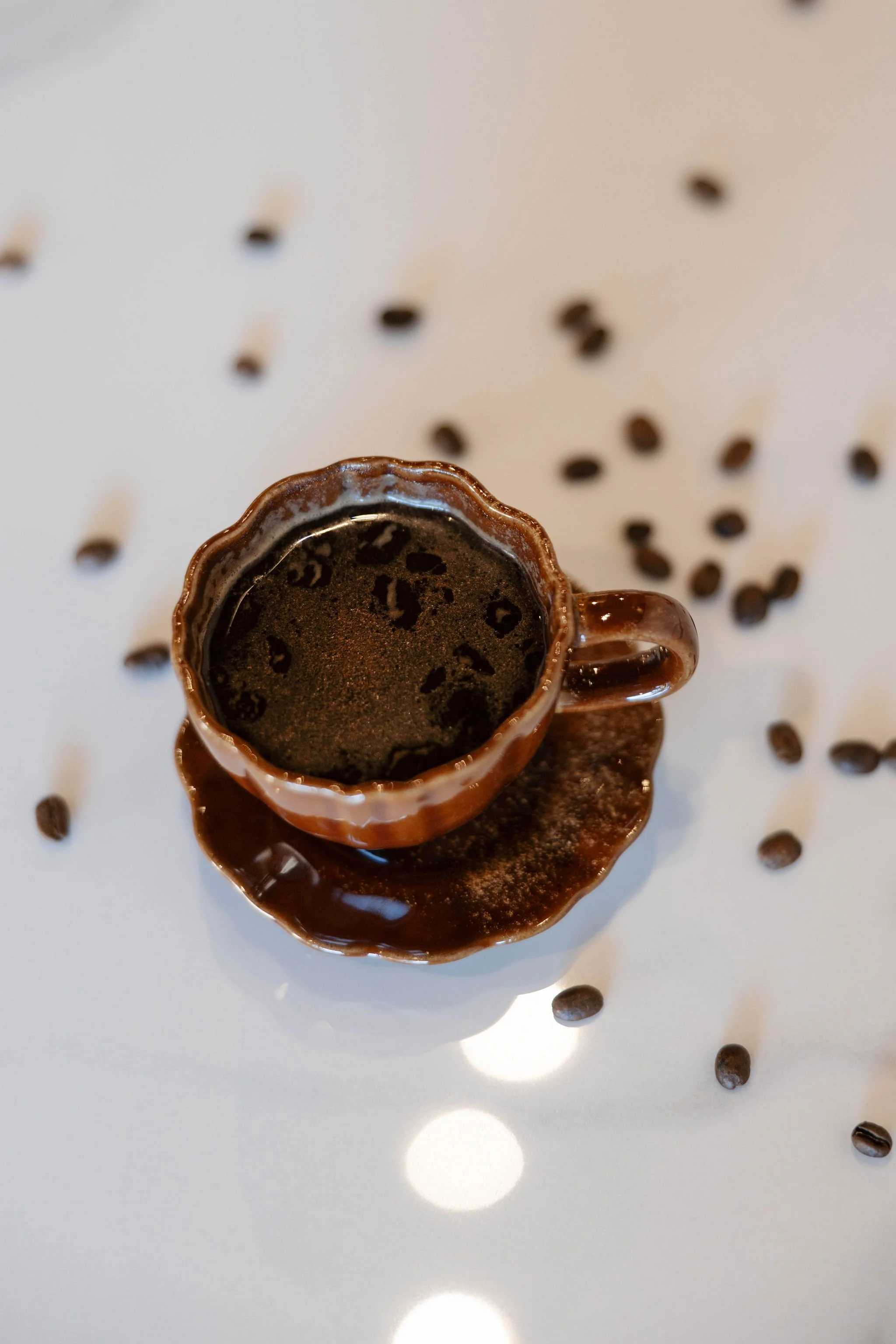 Top-down view of a small hot chocolate in a decorative brown cup and saucer, surrounded by scattered coffee beans on a white surface.