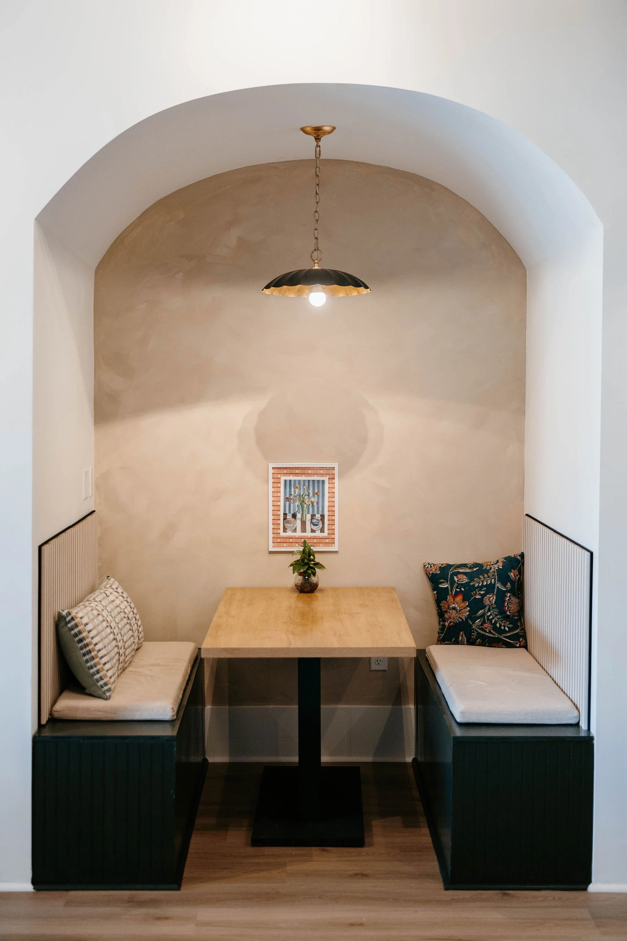 Booth with a wooden table, two benches with cushions, and decorative pillows, behind a small plant, under a hanging light fixture in a cozy nook.