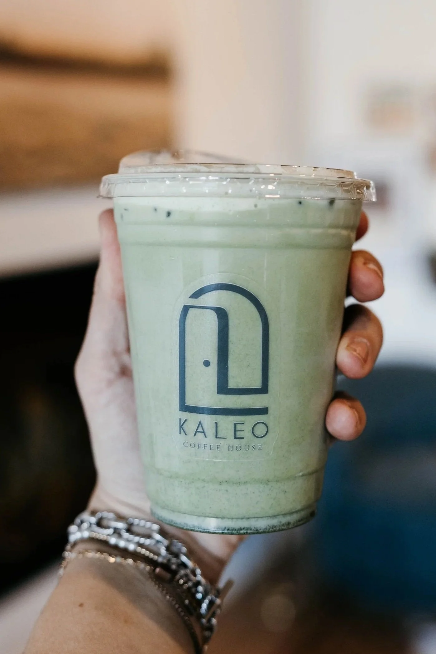 A hand holding a large plastic cup with a clear lid, containing a green blended drink from Kaleo Coffee House.