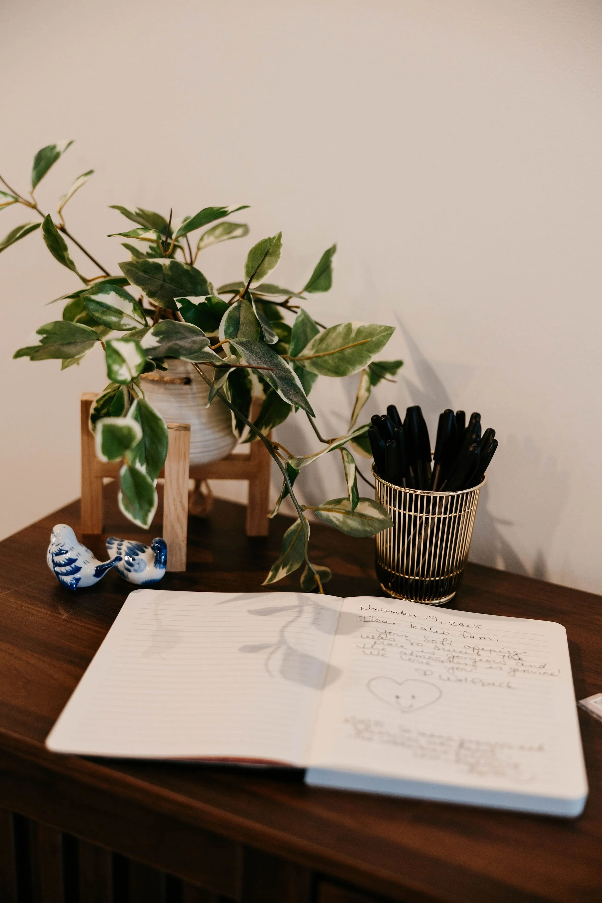 A wooden table with a potted leafy green plant, two small ceramic birds, a gold pen holder with black pens, and an open notebook with handwritten notes and a smiley face drawing.
