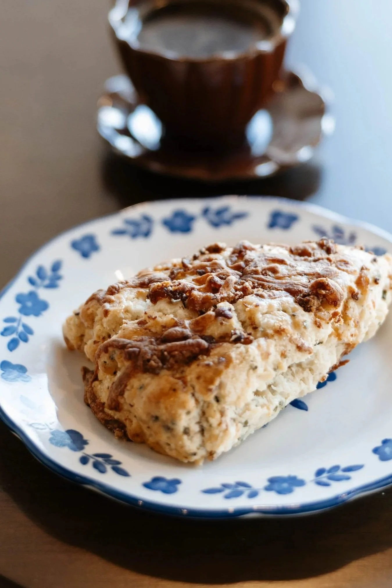 A blueberry muffin on a white plate with blue floral patterns, with a dark coffee mug in the background.