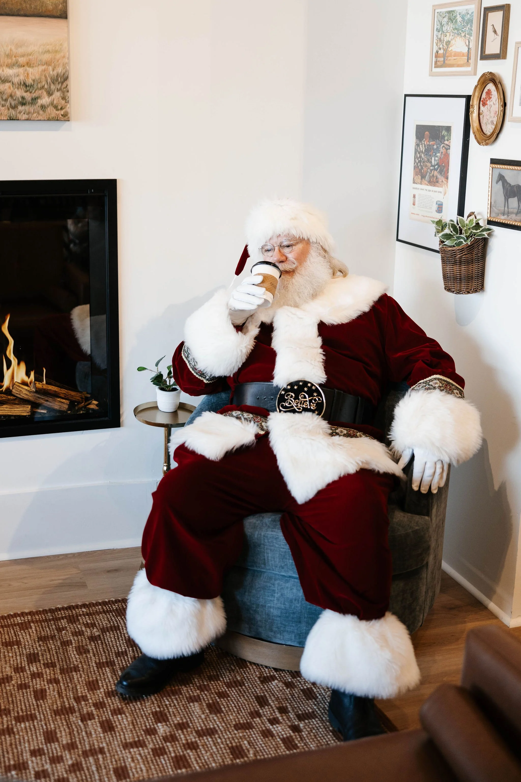Santa Claus sitting in a chair by a fireplace, drinking from a coffee cup, dressed in a traditional red suit with white fur trim, in a decorated living room.