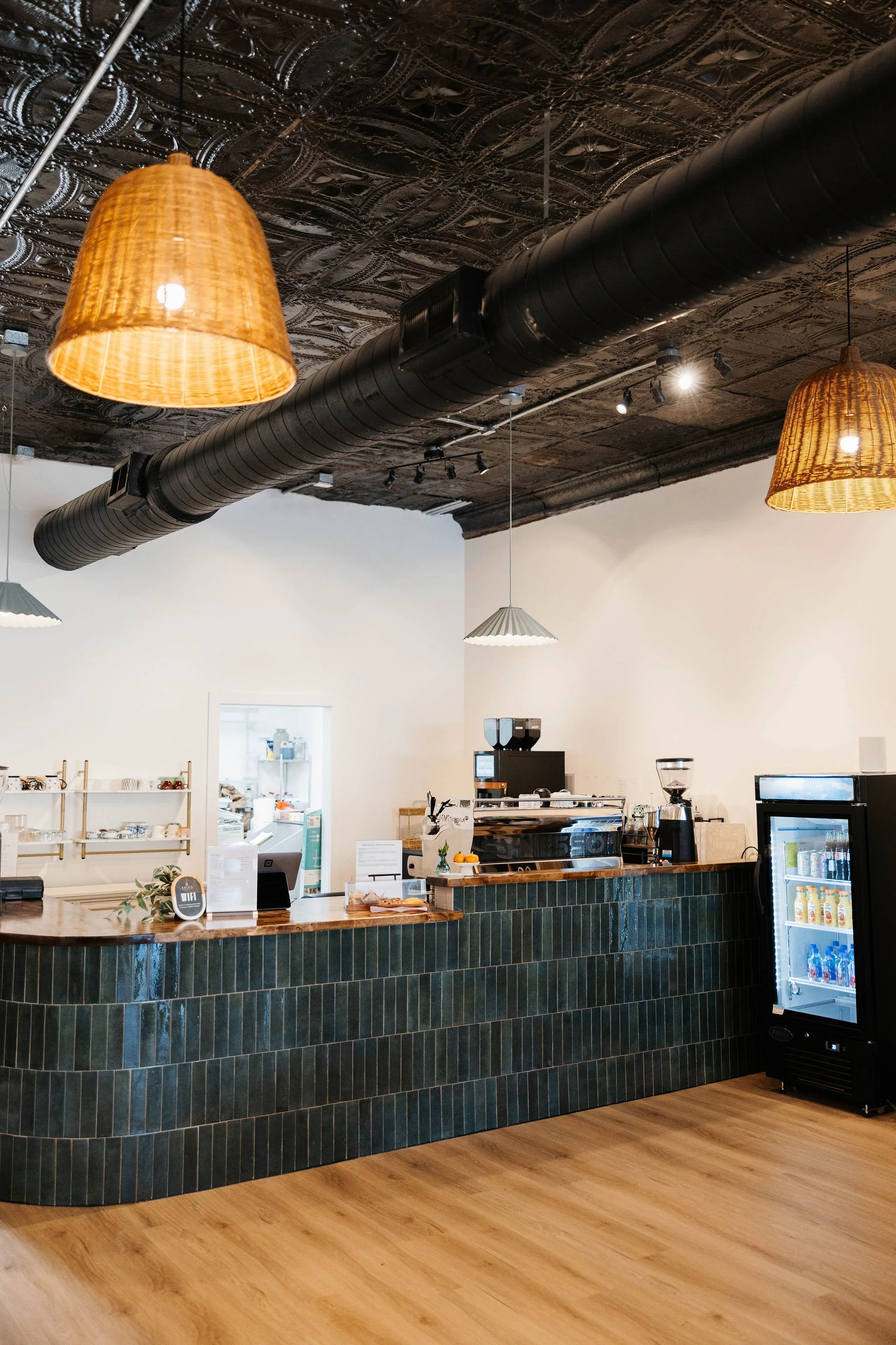 Interior view of a modern cafe with a curved green tiled counter, wooden top, black industrial ductwork on the ornate tin ceiling, hanging wicker lamps, and a beverage fridge.