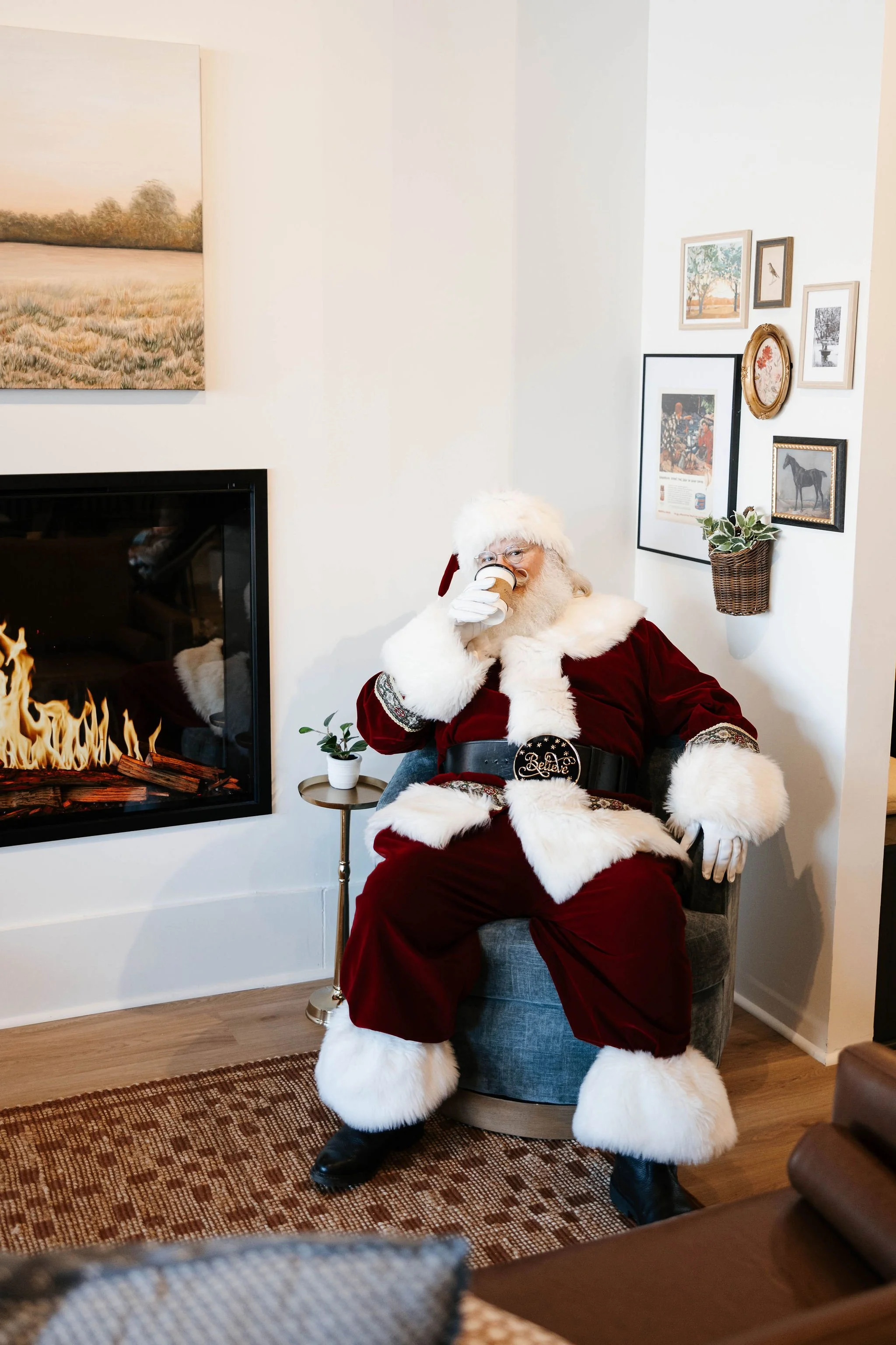Santa Claus sitting in a chair, drinking from a cup, inside a cozy room with a fireplace, framed pictures, and a small plant on a side table.