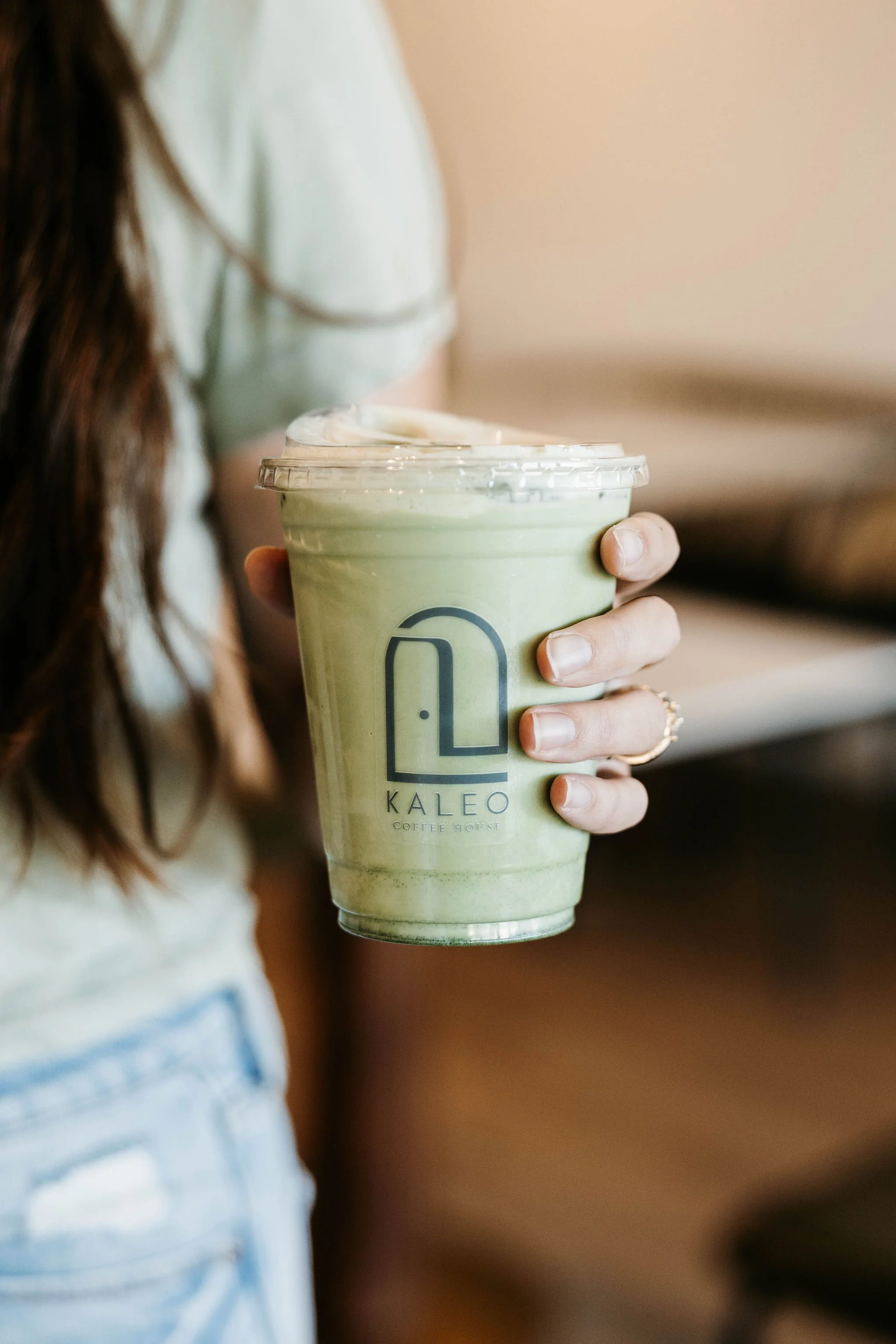 A person holding a large iced matcha latte in a clear plastic cup with a lid, featuring the Kaleo Coffee House logo, with the person's hand and part of their arm visible.