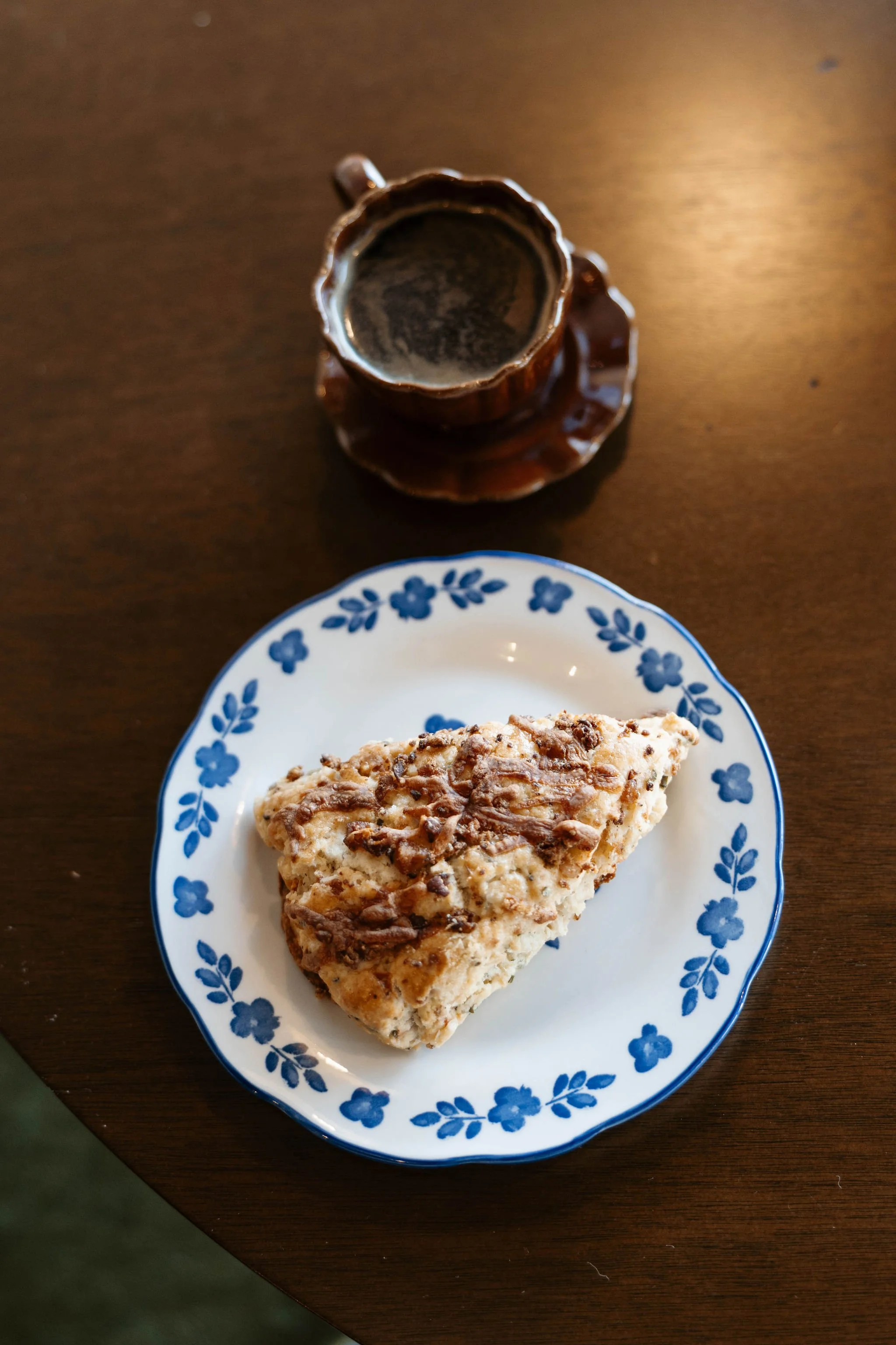 A slice of dessert cake on a white plate with a blue floral pattern, topped with chocolate pieces, on a wooden table. A small brown cup filled with a dark beverage is positioned above the plate.