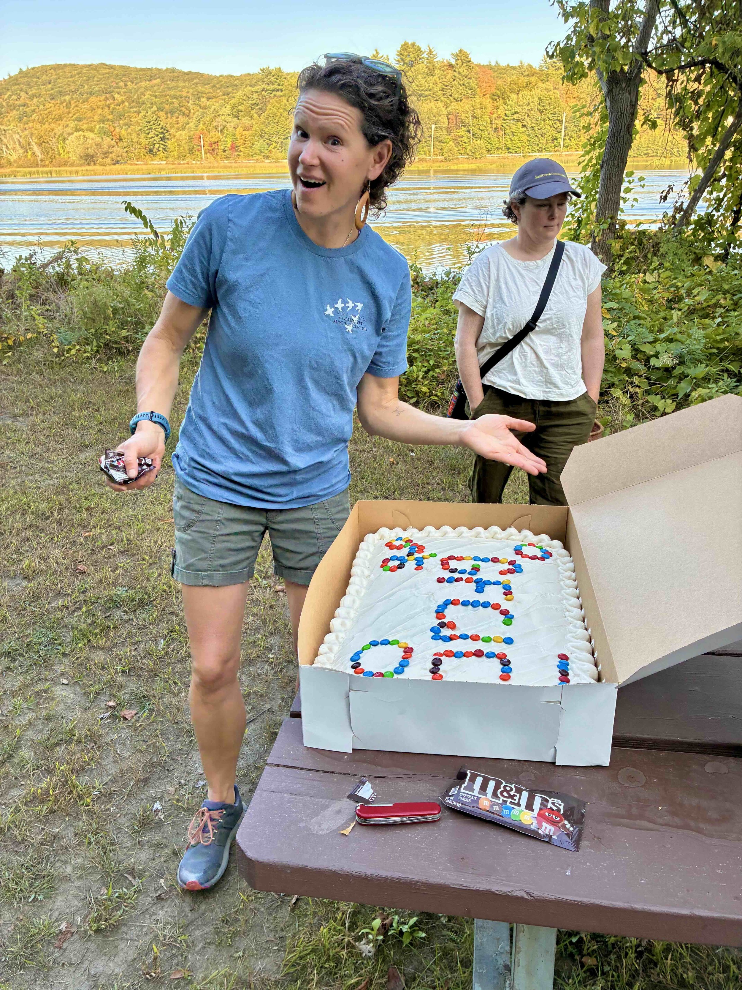 Lauren Beigel MacArthur smiling and gesturing to a large sheet cake that says GJCJC, outdoors at a picnic table with a lake in the background