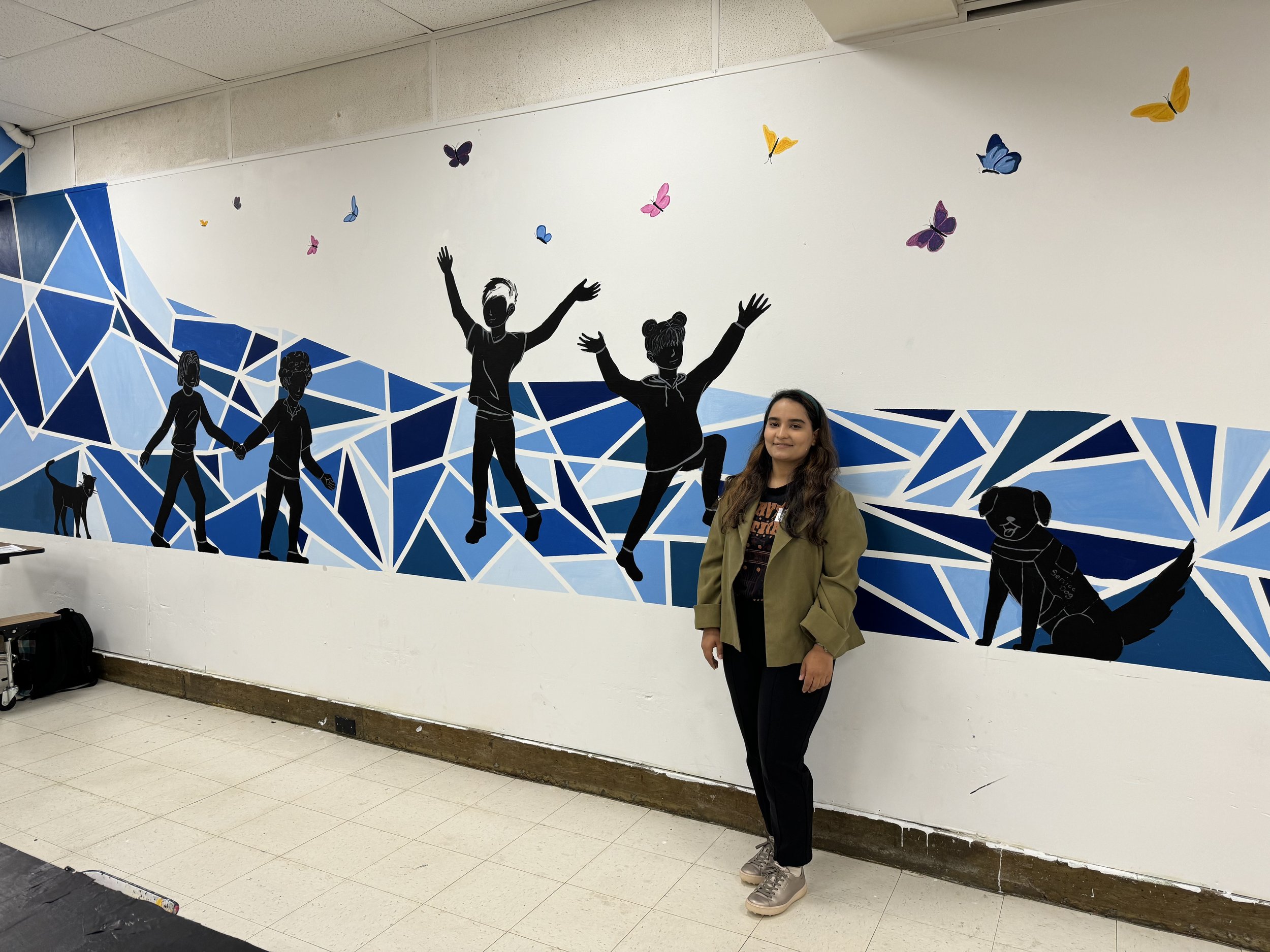 Negina Azimi standing in front of a wall with a mural composed of geometric shapes in various shades of blue, two figures of people holding hands, two figures with arms raised, a cat, a dog, and butterflies