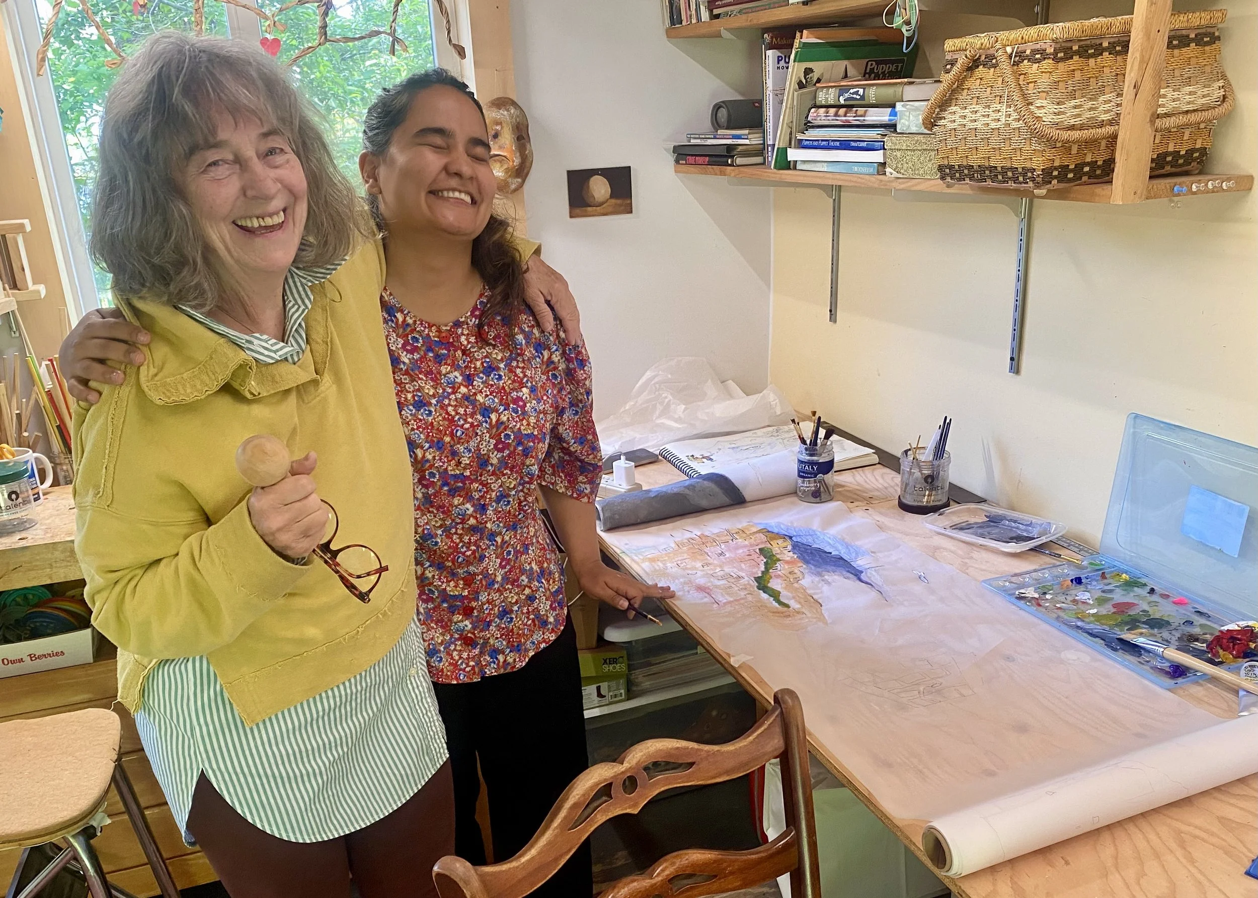 Jana Zeller Bass and Negina Azimi with arms around each other laughing next to a table with a rolled scroll and a shelf with a basket and books
