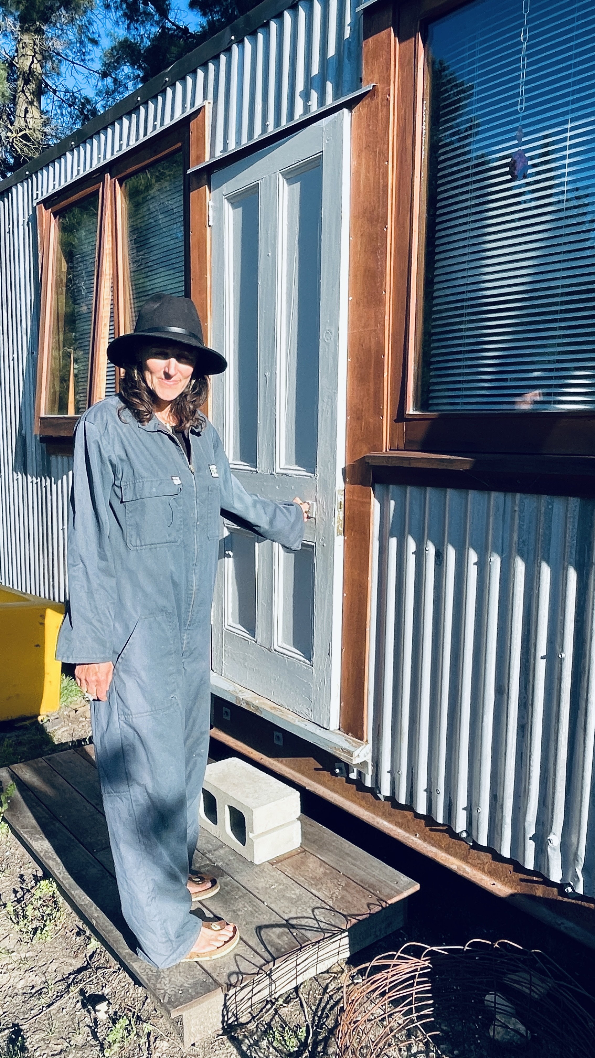 Woman in blue jumpsuit and black hat standing on wooden porch in front of a teal door with her hand on door handle, beside a building with corrugated metal siding and a window with blinds.