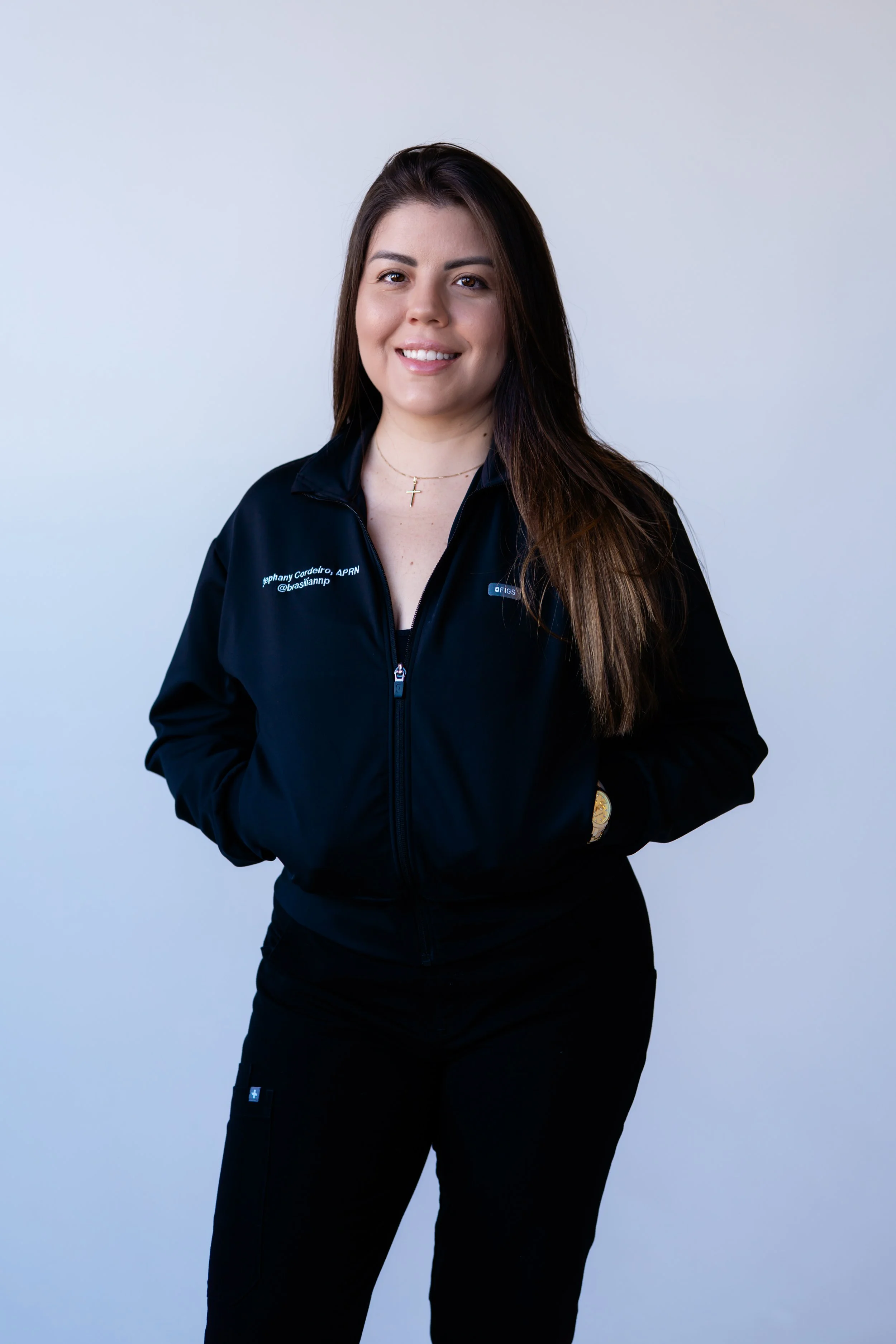 A young woman with long brown hair smiling, wearing a black jacket and black pants, standing against a plain light gray background.