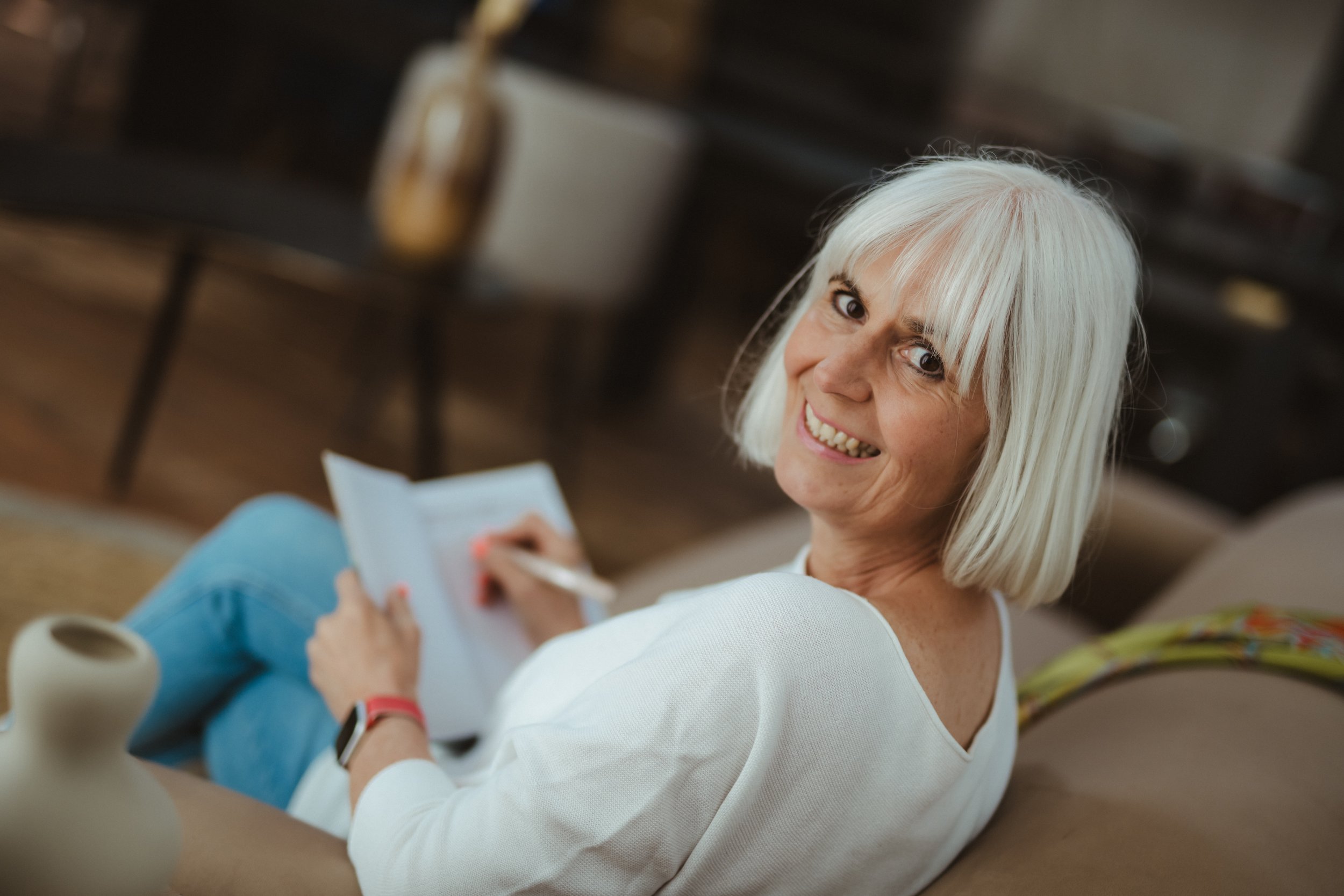 Eine ältere Frau mit weißem Haar sitzt auf einem Sofa, lächelt in die Kamera und schreibt in ein Notizbuch.