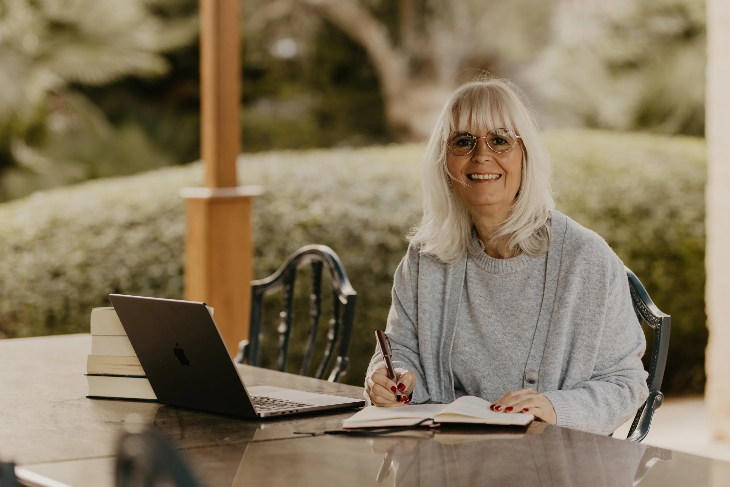 Eine ältere Frau mit weißen Haaren und Brille sitzt an einem Holztisch im Freien, hebt einen Kugelschreiber und blickt lächelnd in die Kamera. Auf dem Tisch liegt ein offenes Notizbuch, ein Laptop und einige Bücher, im Hintergrund ist eine grüne, bewaldete Landschaft.