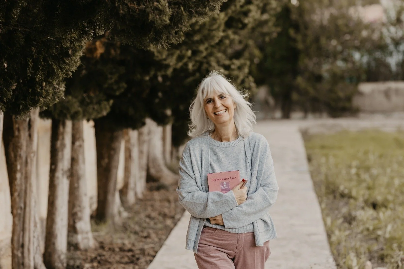Eine lachende ältere Frau mit weißen Haaren, die einen roten Gedichtband an der Brust hält, steht auf einem Gehweg neben einem Baumreihe in einer Parklandschaft