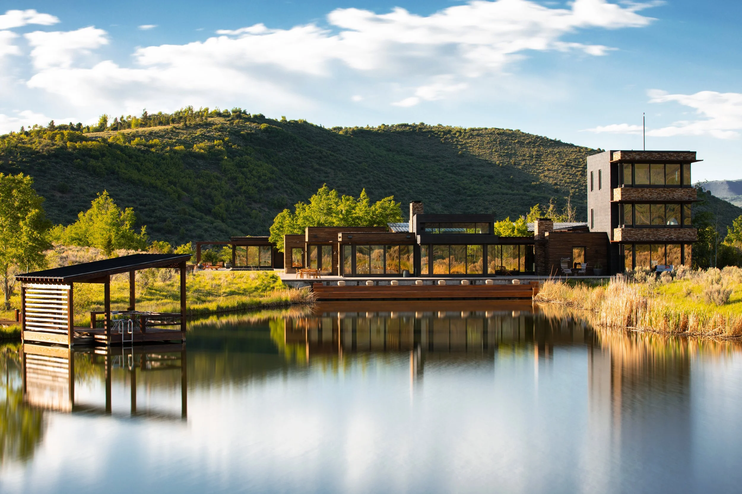Modern house with large glass windows, a reflecting pond in front, surrounded by greenery and hills under a partly cloudy sky.