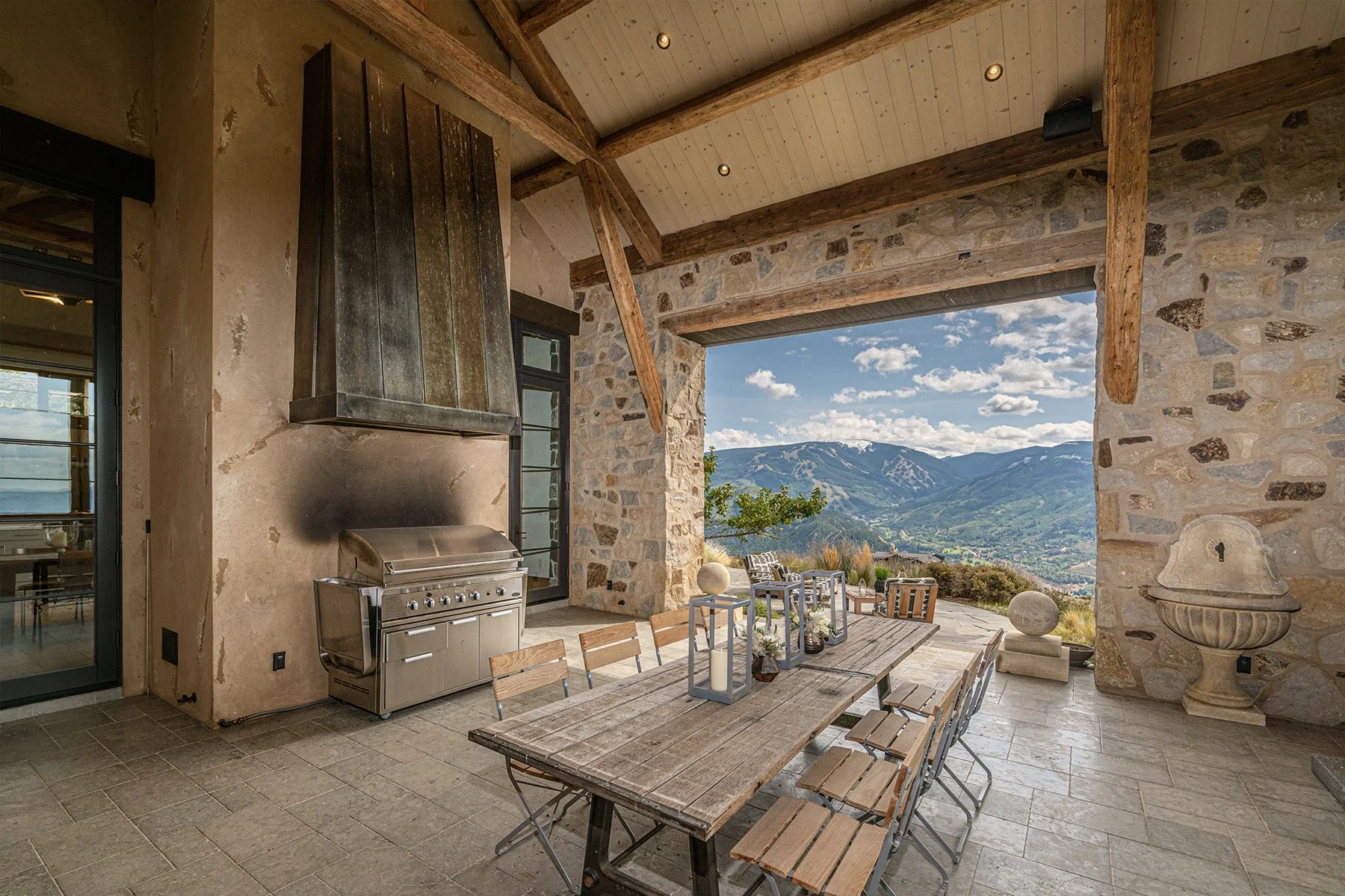 Covered outdoor patio with a rustic wooden dining table, matching chairs, and decorative candle lanterns, overlooking mountains and a valley with trees and hills.