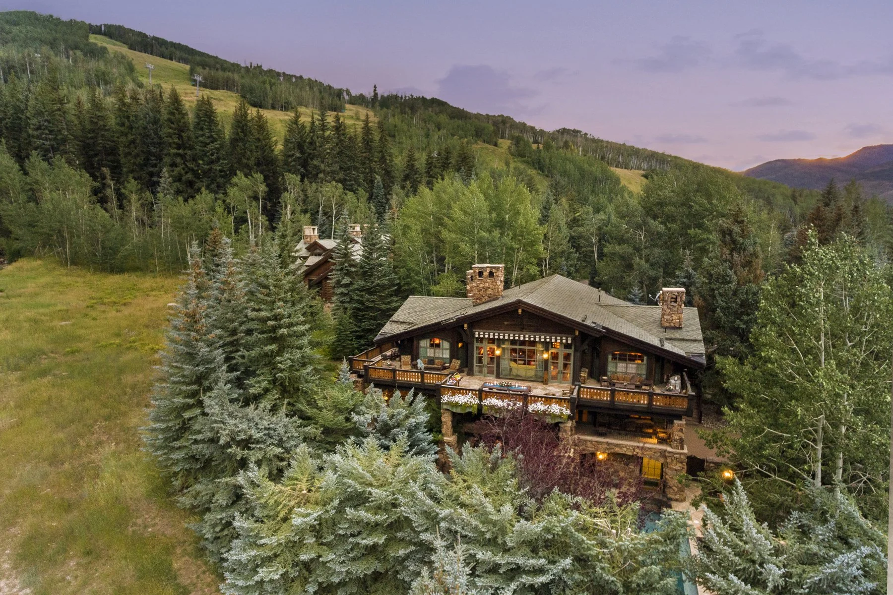 A large house built in a mountain landscape surrounded by tall trees, with a balcony and outdoor lighting at sunset.