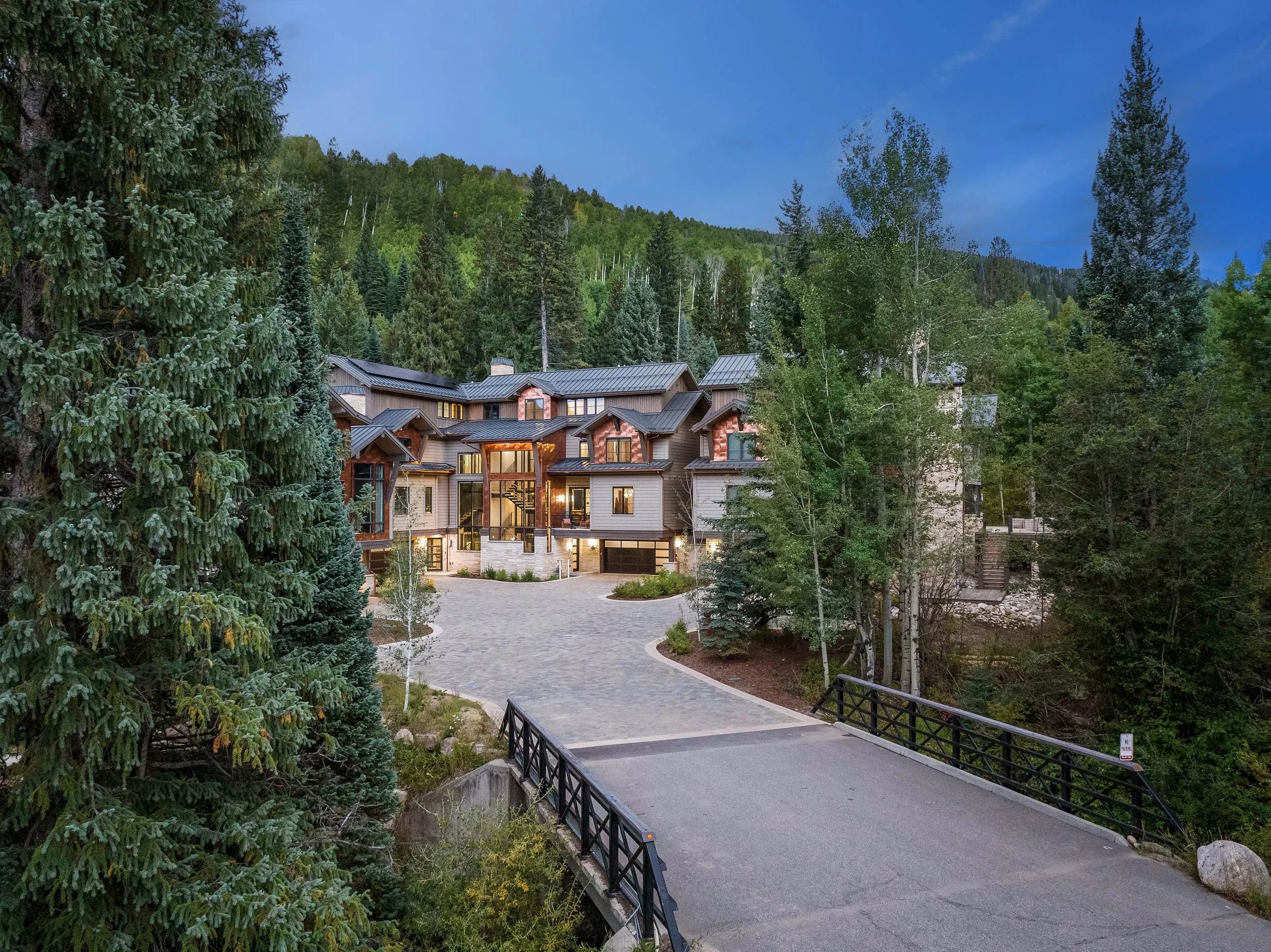 Large modern house with multiple levels surrounded by forest, seen from the driveway with a small bridge in the foreground.