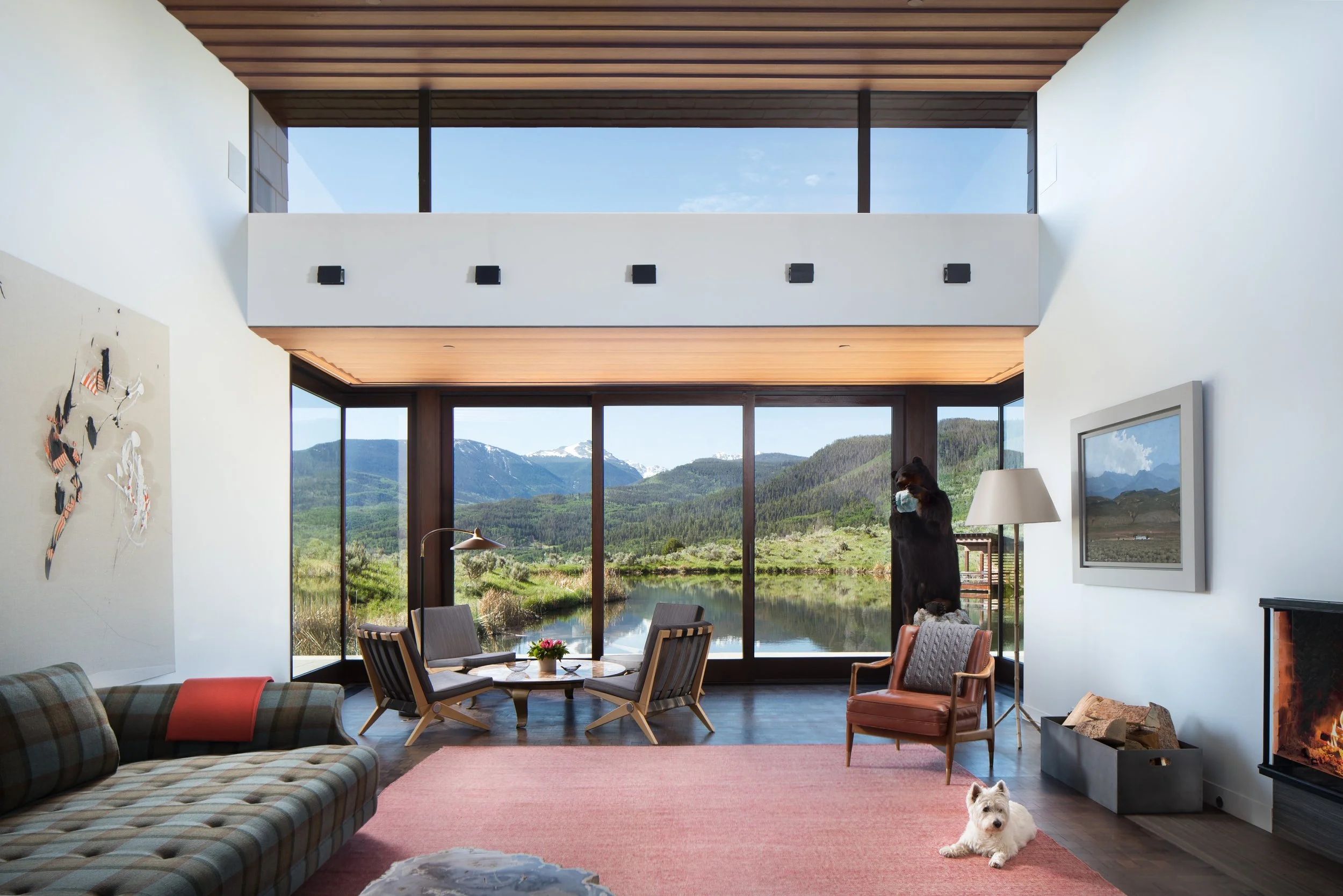 Interior of a modern living room with large floor-to-ceiling windows revealing a mountain river landscape, featuring a plaid sofa, a pink area rug, a white dog lying on the rug, a vintage armchair, a floor lamp, wall art, a picture frame, and a fireplace.