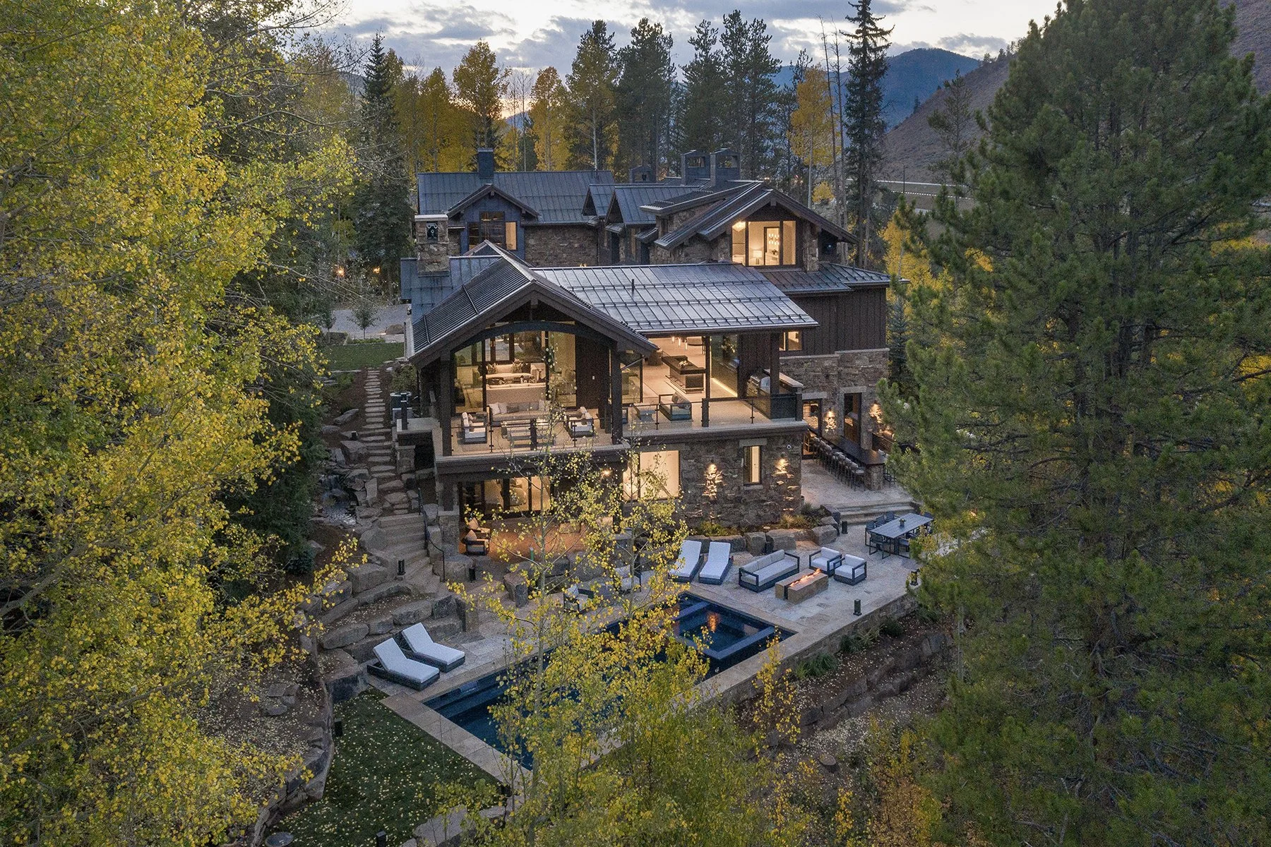 A large modern house with multiple levels, surrounded by trees and mountains, illuminated at dusk. It features a pool area with lounge chairs and an outdoor dining space.