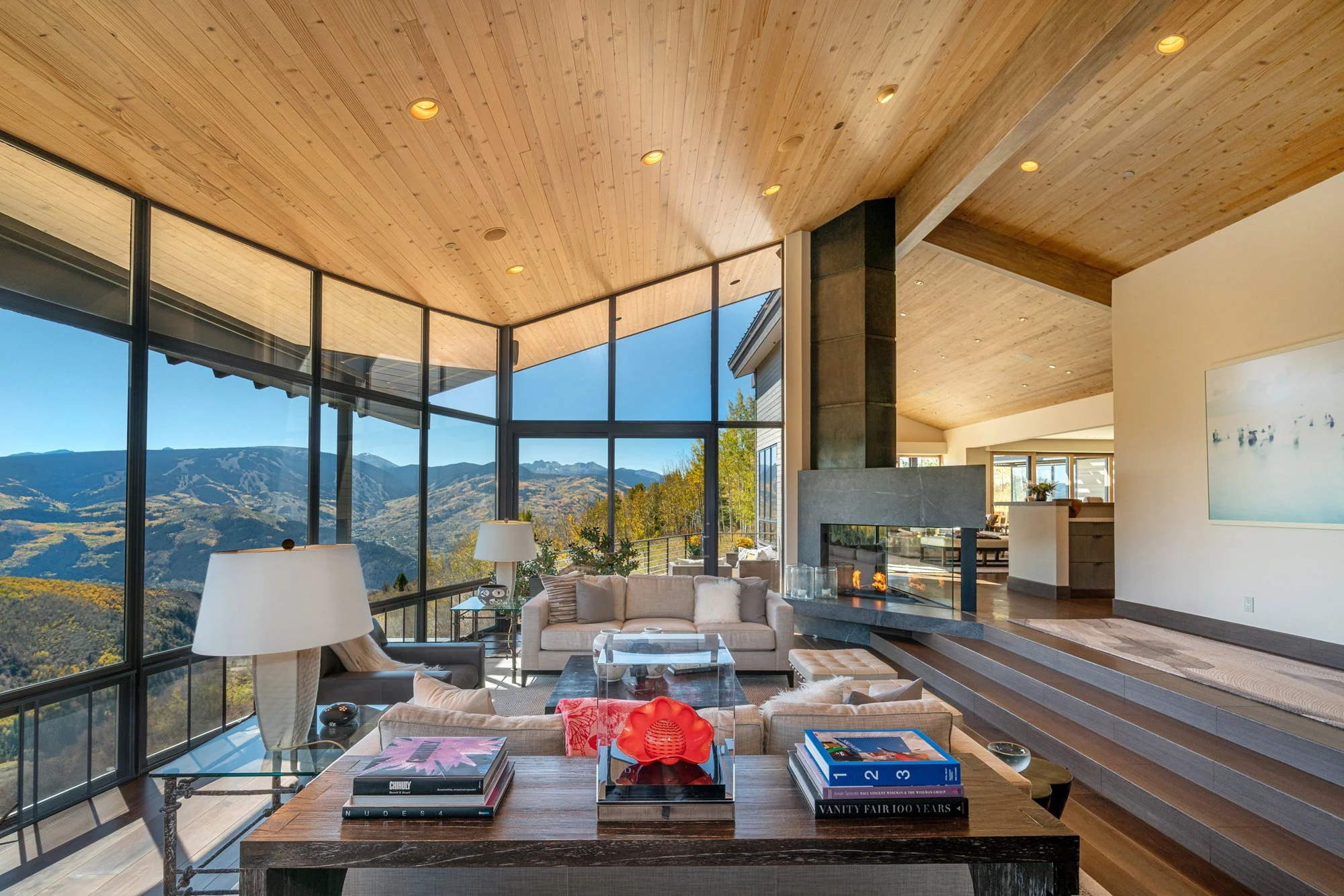 Modern living room with large glass windows overlooking scenic mountain landscape, featuring beige sofas, coffee tables, lamps, a fireplace, and wooden ceiling.