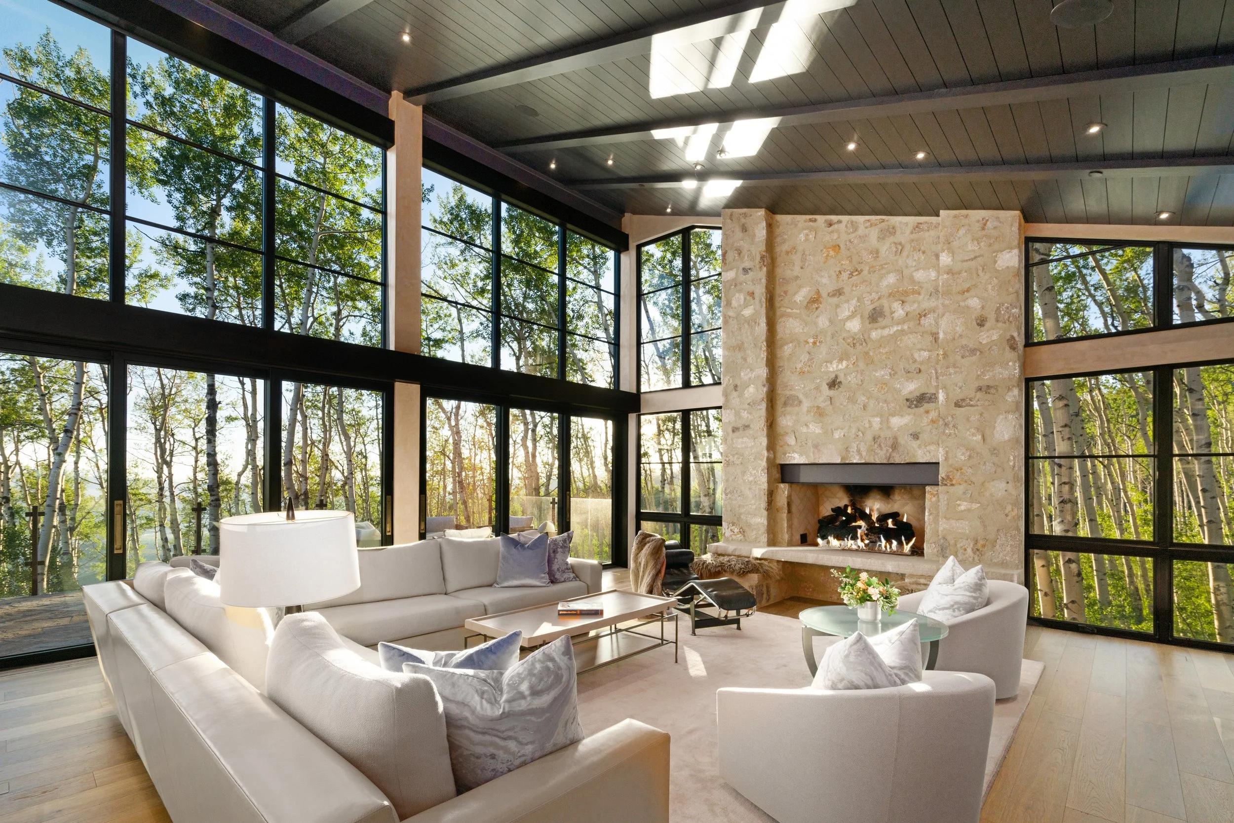 Living room with large floor-to-ceiling windows showing a forest view, a stone fireplace, and modern white furniture including a sectional sofa, armchairs, and a glass coffee table.