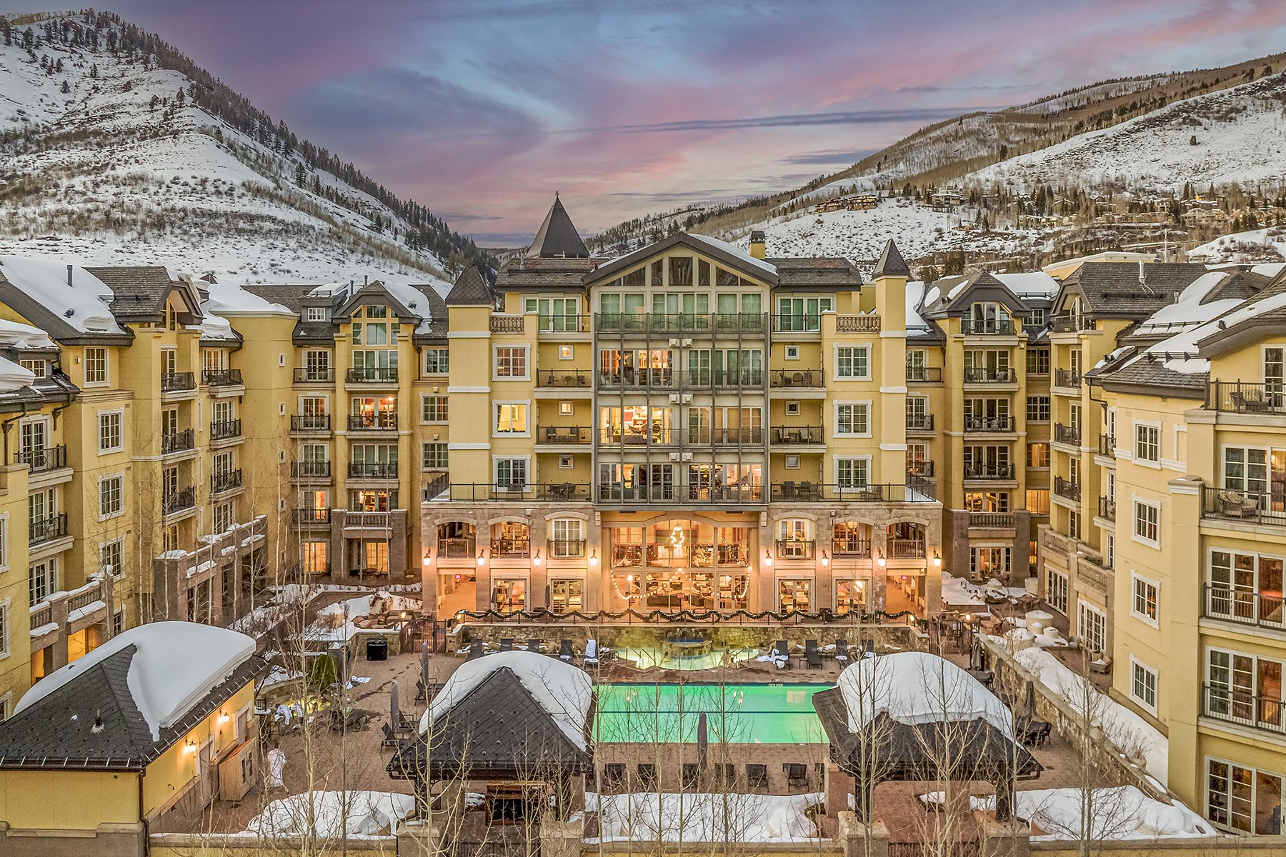 A multi-story mountain resort with yellow buildings surrounding a heated outdoor swimming pool, with snow-covered hills in the background at sunset.