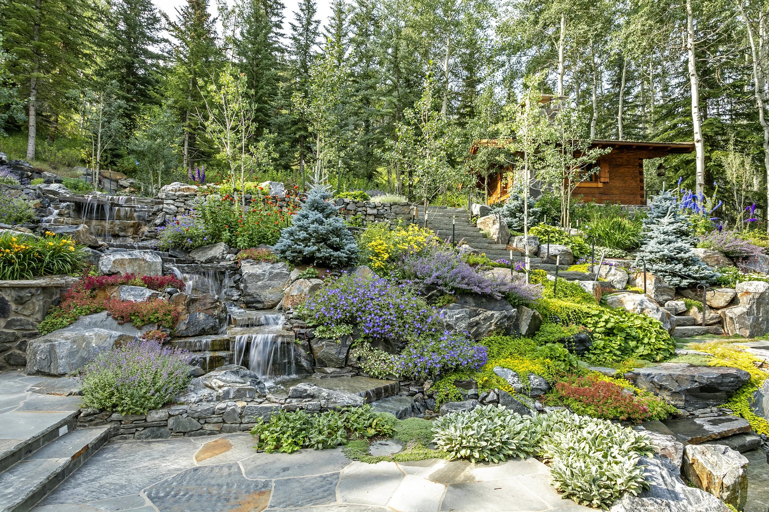 A lush garden landscape with a cascading waterfall feature, surrounded by colorful flowers, shrubs, and trees, with a wooden shed in the background.