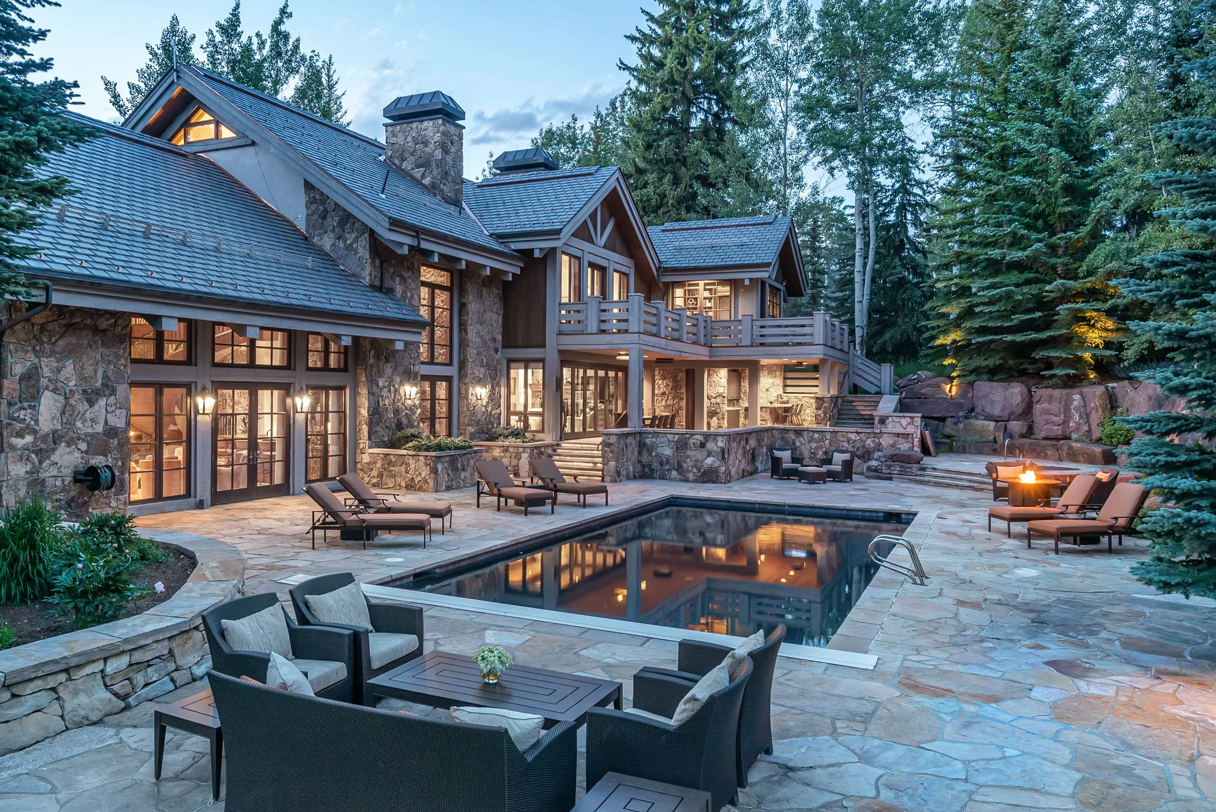 A luxurious house with a stone exterior, large windows, and wooden balconies, surrounded by trees, with a swimming pool and outdoor seating area in the backyard during evening.