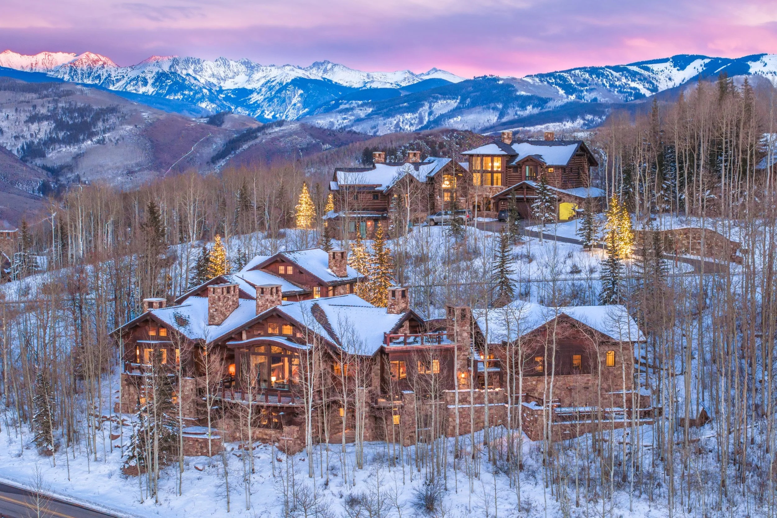 A mountain retreat with large wooden houses, snow-covered rooftops, and lit-up Christmas trees on a hillside with snow and deciduous trees, with snow-capped mountains in the background at dusk.