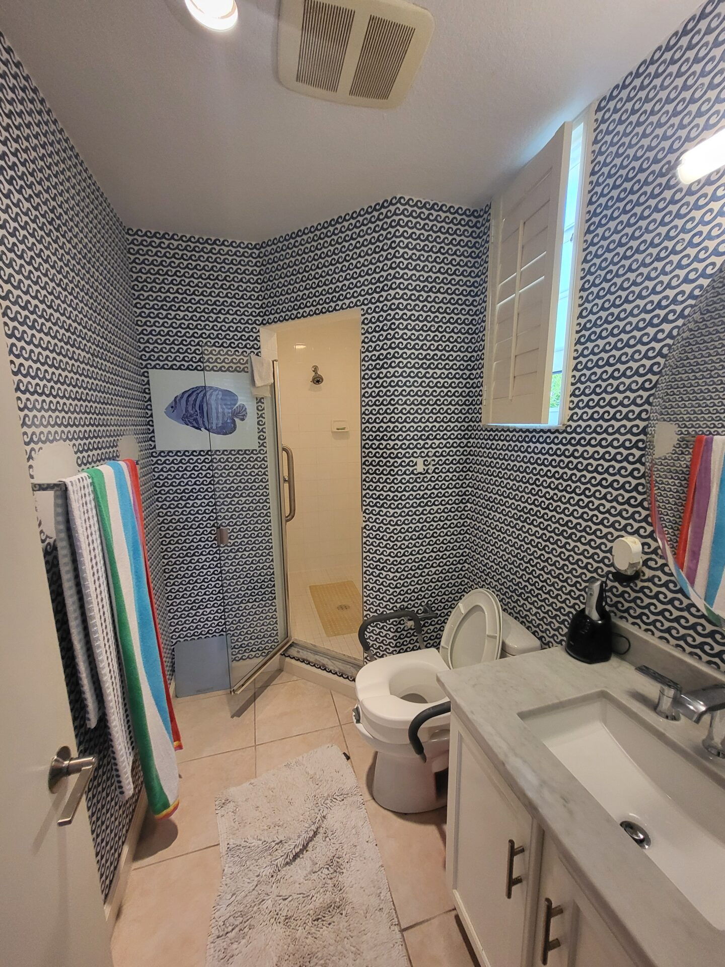 Bathroom with blue wave-patterned wallpaper, a small shower with glass door, a toilet with seat raised, a white vanity with marble top, a black soap dispenser, a mirror, a window with plantation shutters, striped towels hanging, and a beige bath mat on tiled floor.