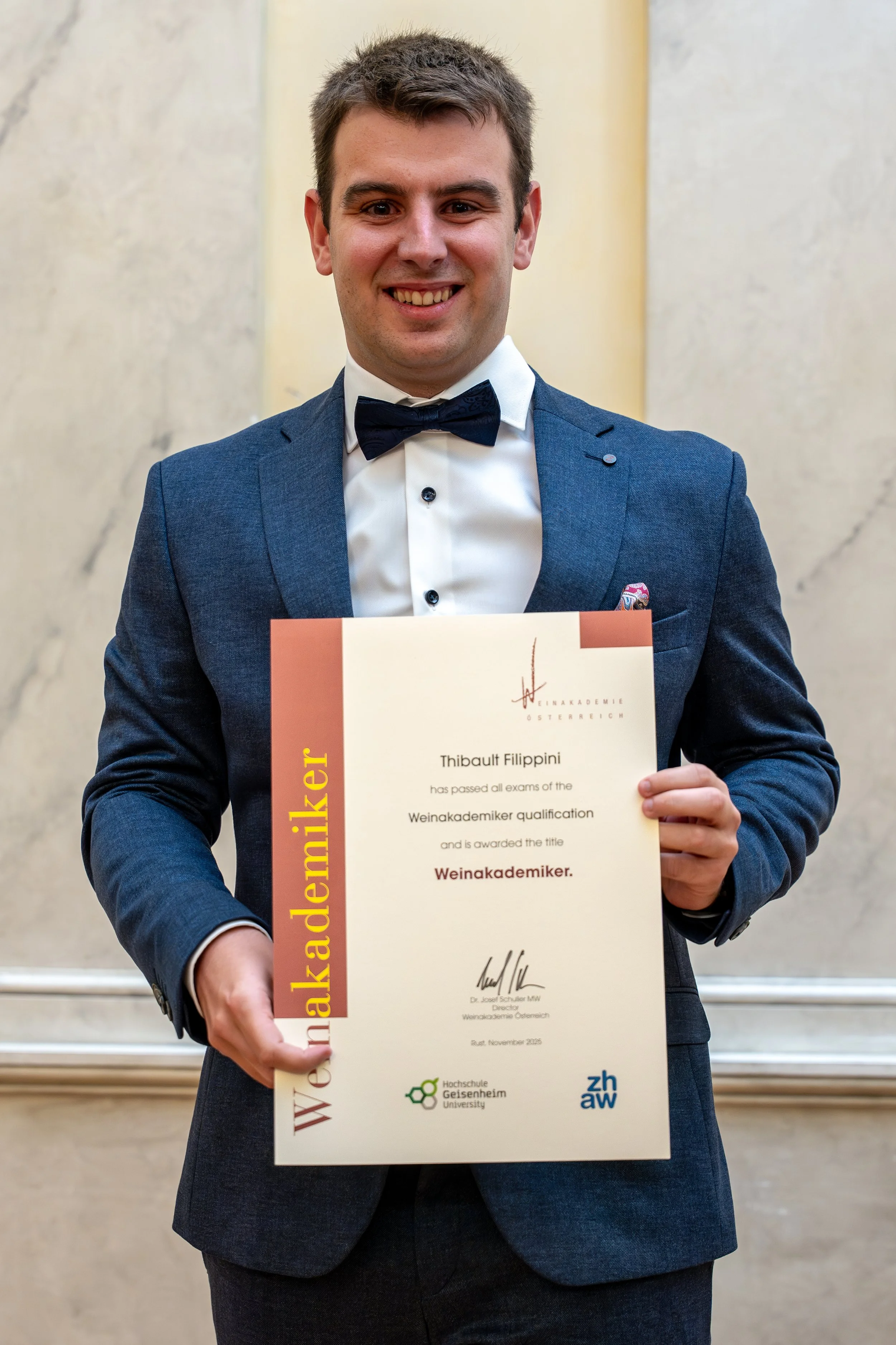 Portretfoto van Thibault Filippini die trots zijn Weinakademiker (DipWSET) diploma vasthoudt, glimlachend naar de camera. Hij staat in een lichte, professionele omgeving, wat zijn expertise en toewijding aan wijneducatie benadrukt